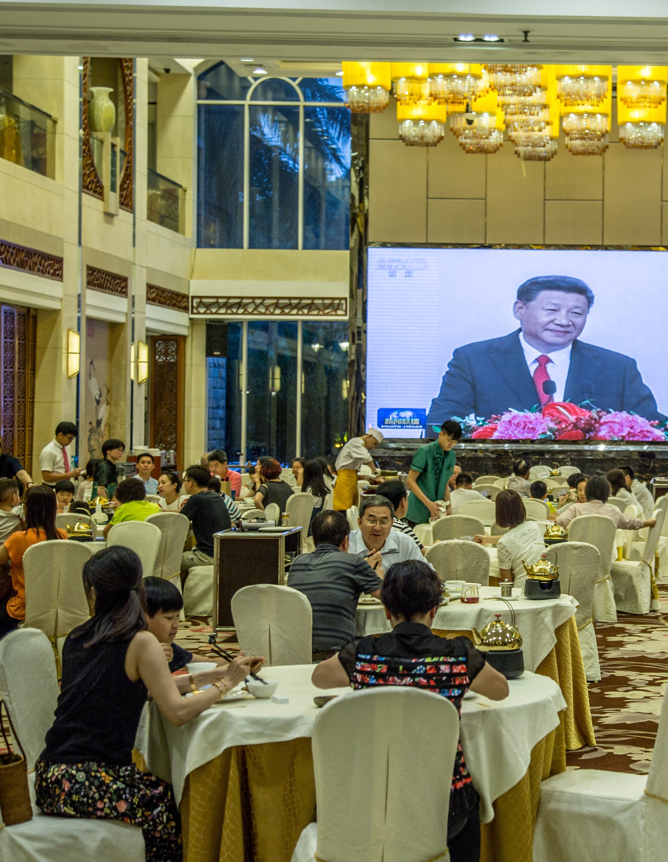 Grand banquet hall filled with diners at round tables beneath ornate chandeliers with a large projection screen broadcasting at the front