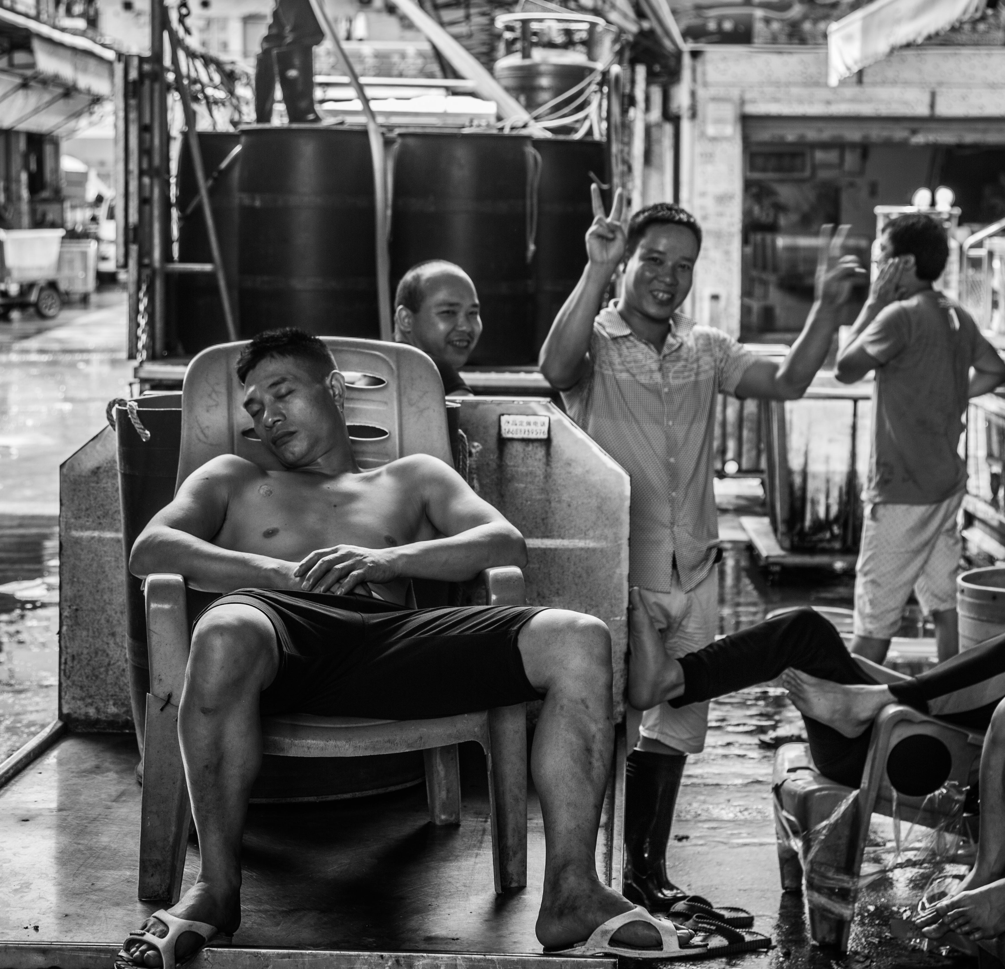 Black and white photo of a grinning fish market worker flashing a peace sign while his shirtless colleague sleeps in a plastic chair