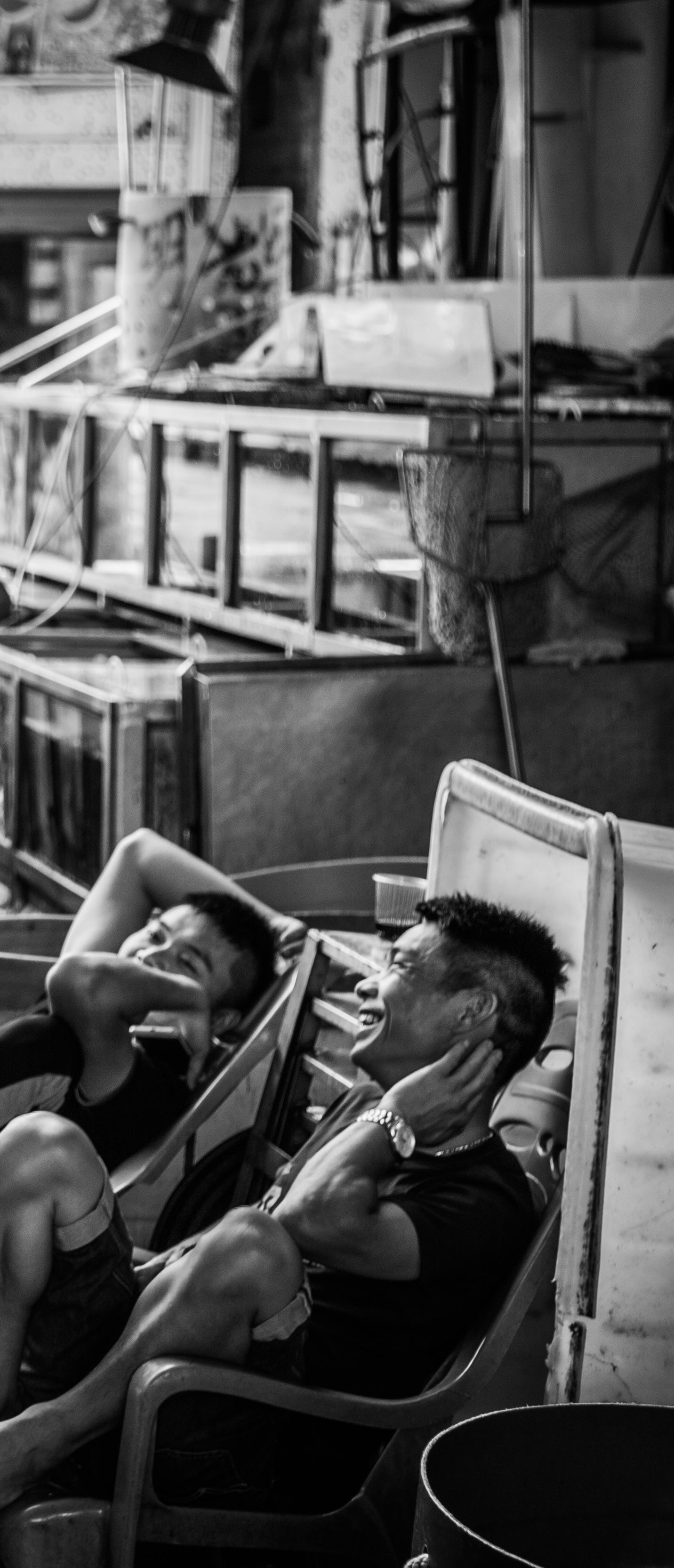 Black and white vertical image of two fish market workers laughing while resting in plastic chairs among empty glass tanks and equipment