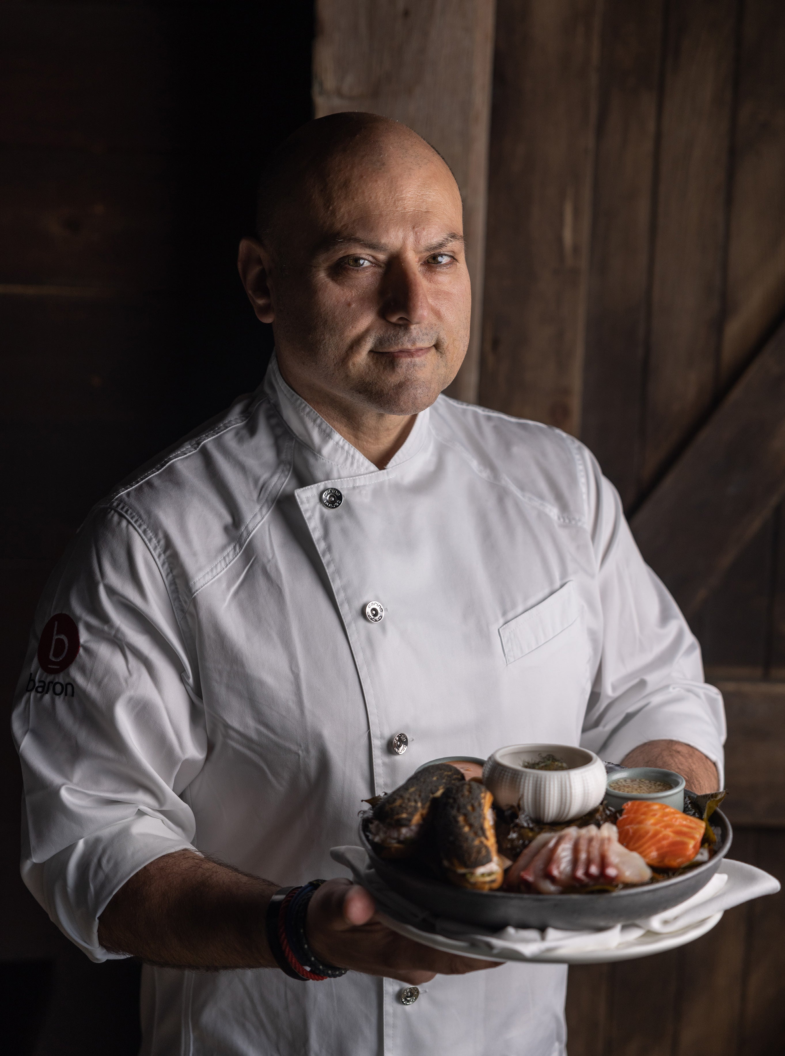 Bald chef in white chef jacket holding a platter of oysters and seafood, lit by soft window light against a dark wooden background