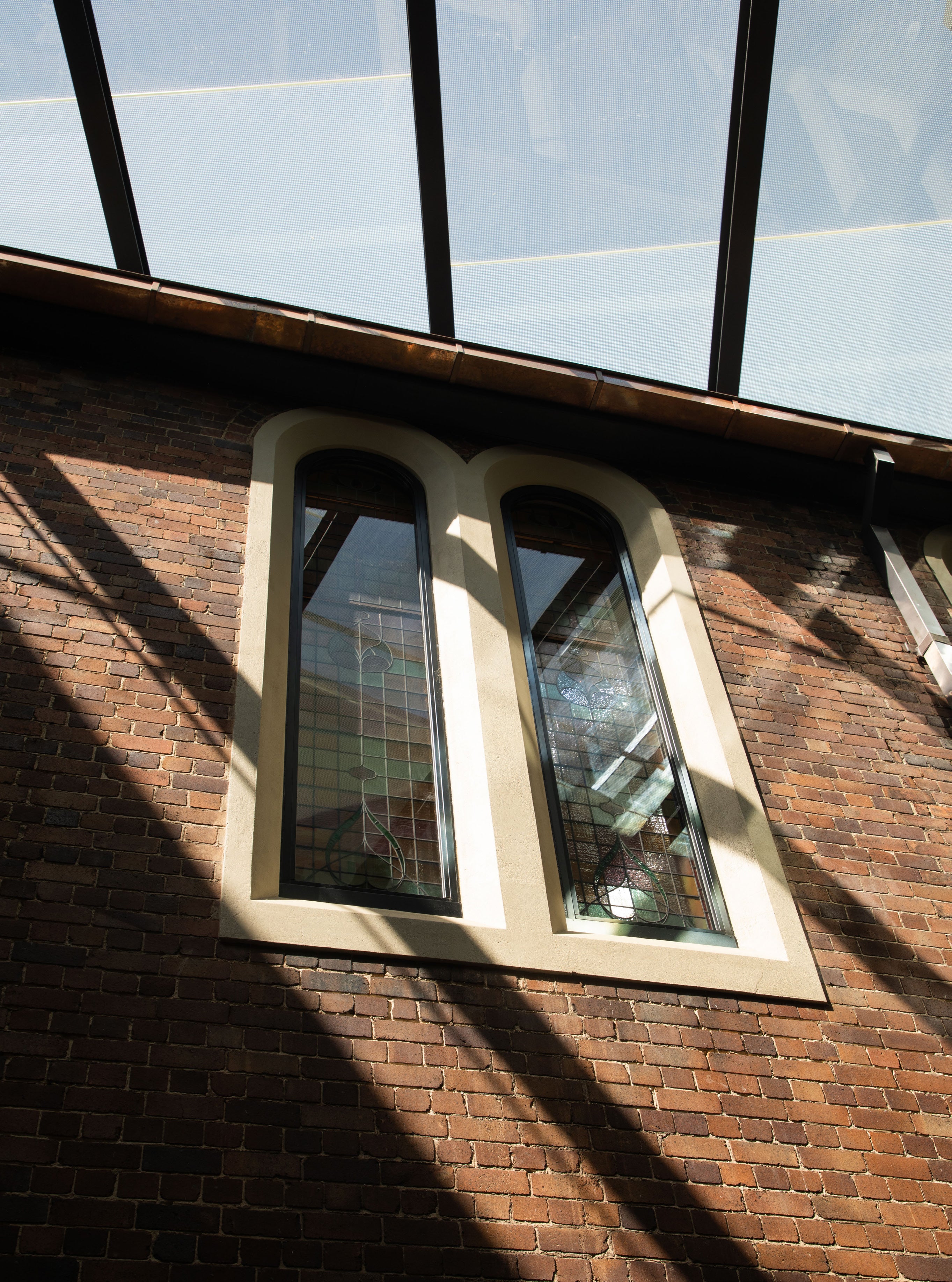 Twin arched windows set into a red brick wall beneath a glass skylight casting diagonal shadows across the facade