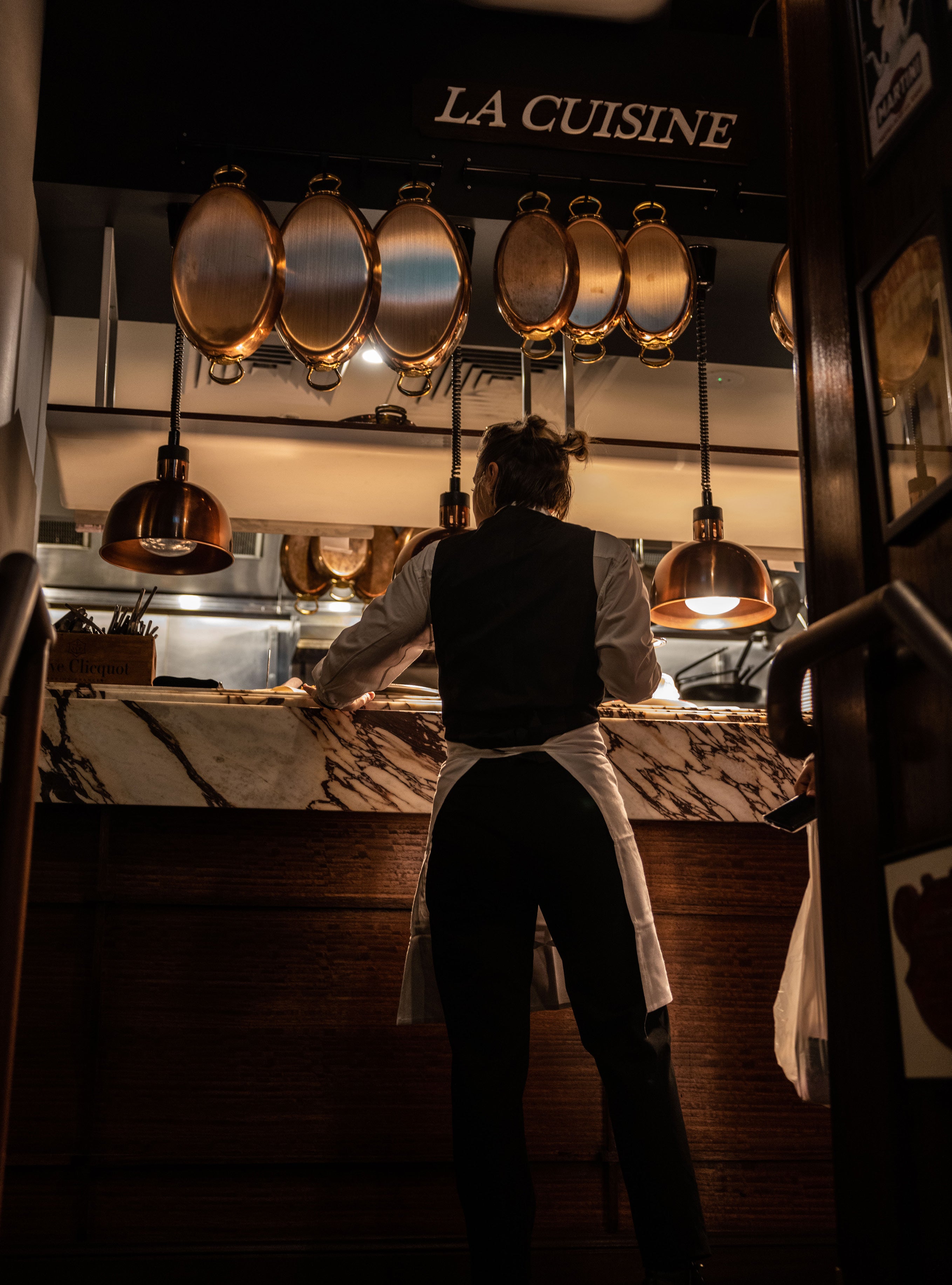 Silhouetted waiter in vest and apron standing at a marble kitchen pass beneath hanging copper pans and warm pendant lights with La Cuisine sign above