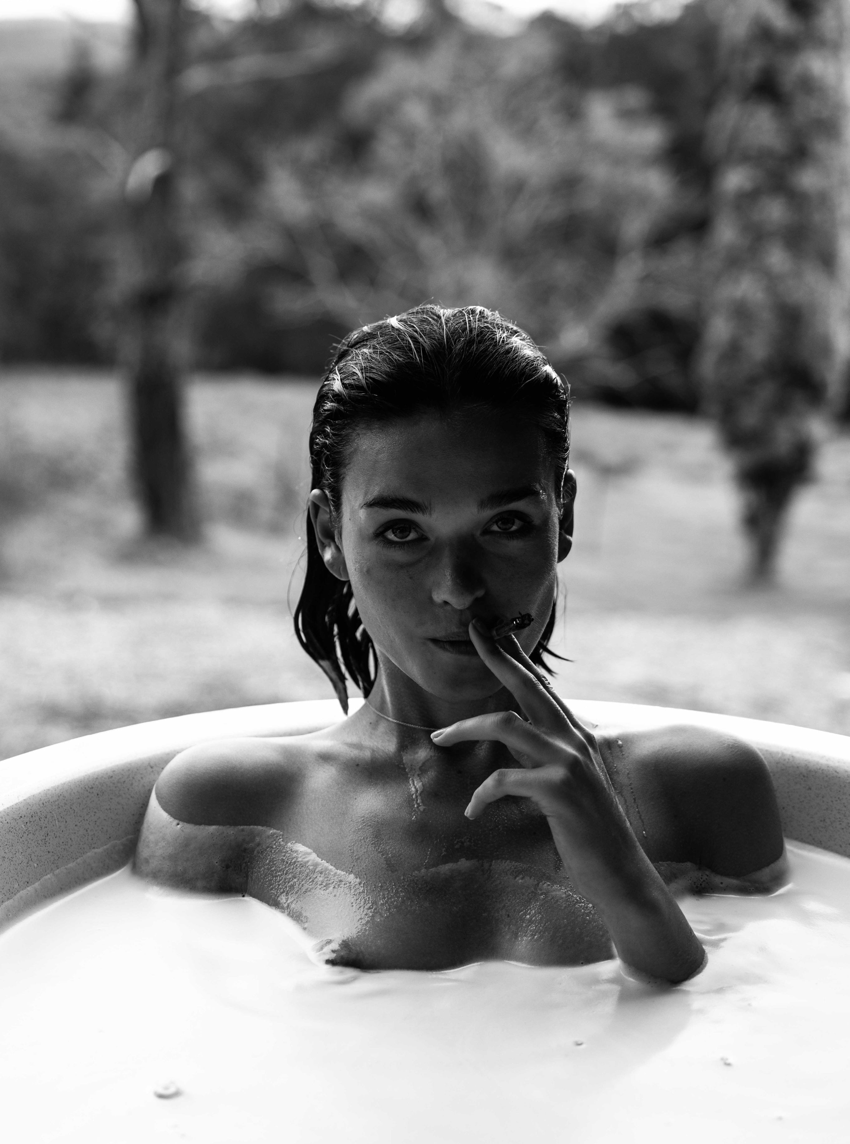 Black and white portrait of a woman with wet slicked-back hair in a bath looking directly at camera with a finger touching her lips