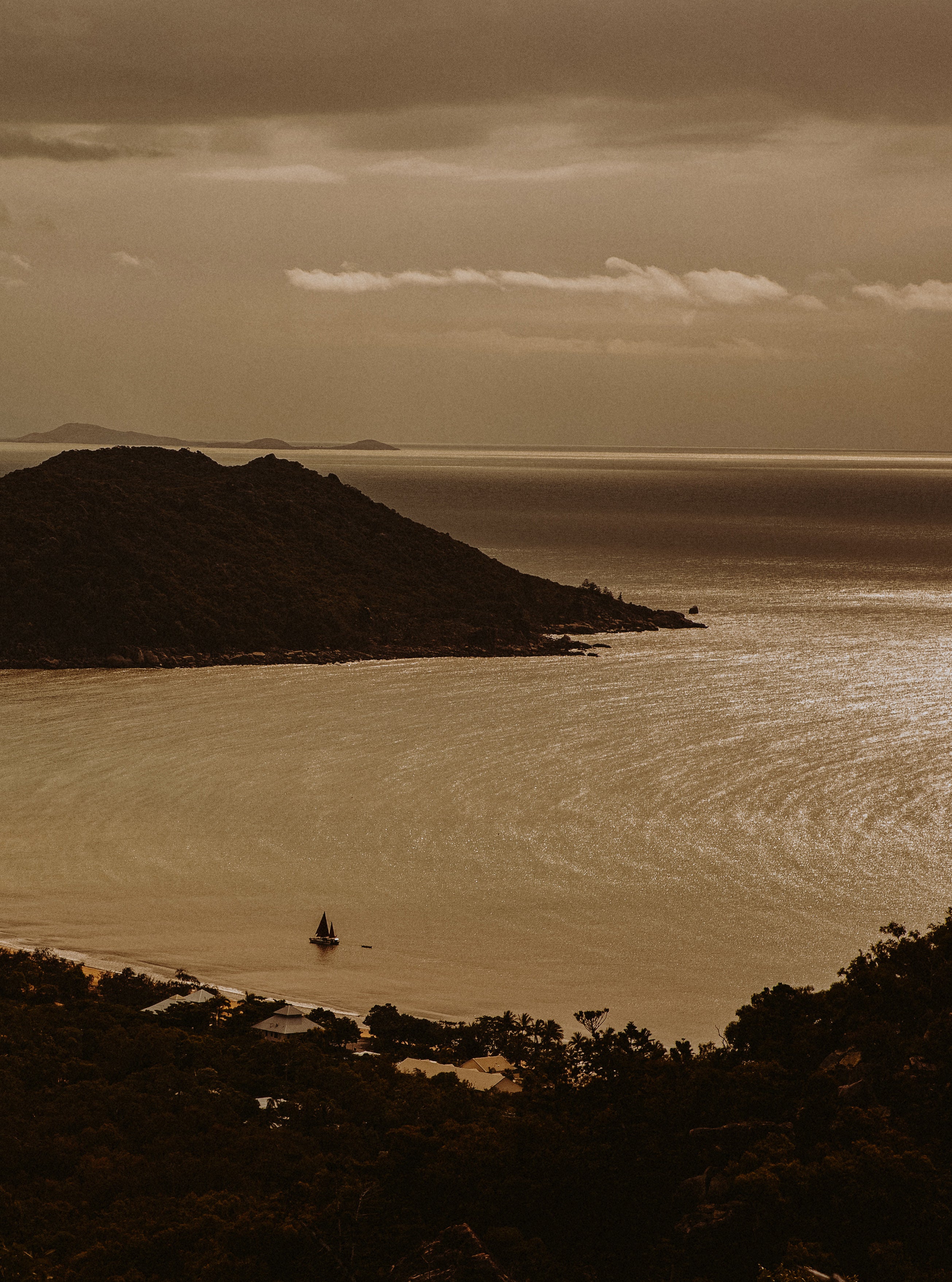 Sepia-toned aerial view of a calm bay at dusk with a single sailboat, silhouetted headland and dramatic cloudy sky