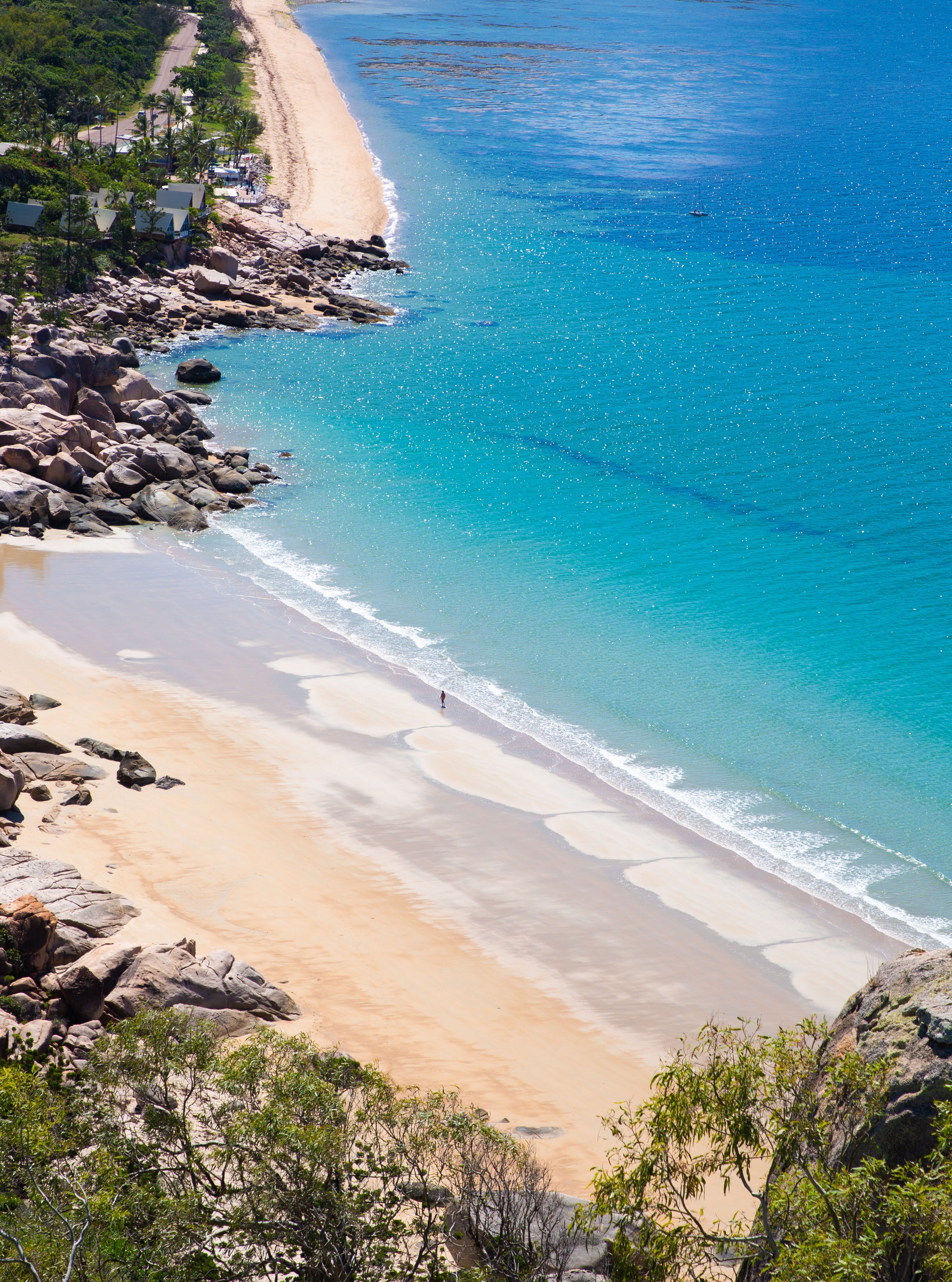 Elevated aerial view of a curved tropical beach with golden sand bordered by granite boulders and vivid turquoise water with two small figures walking