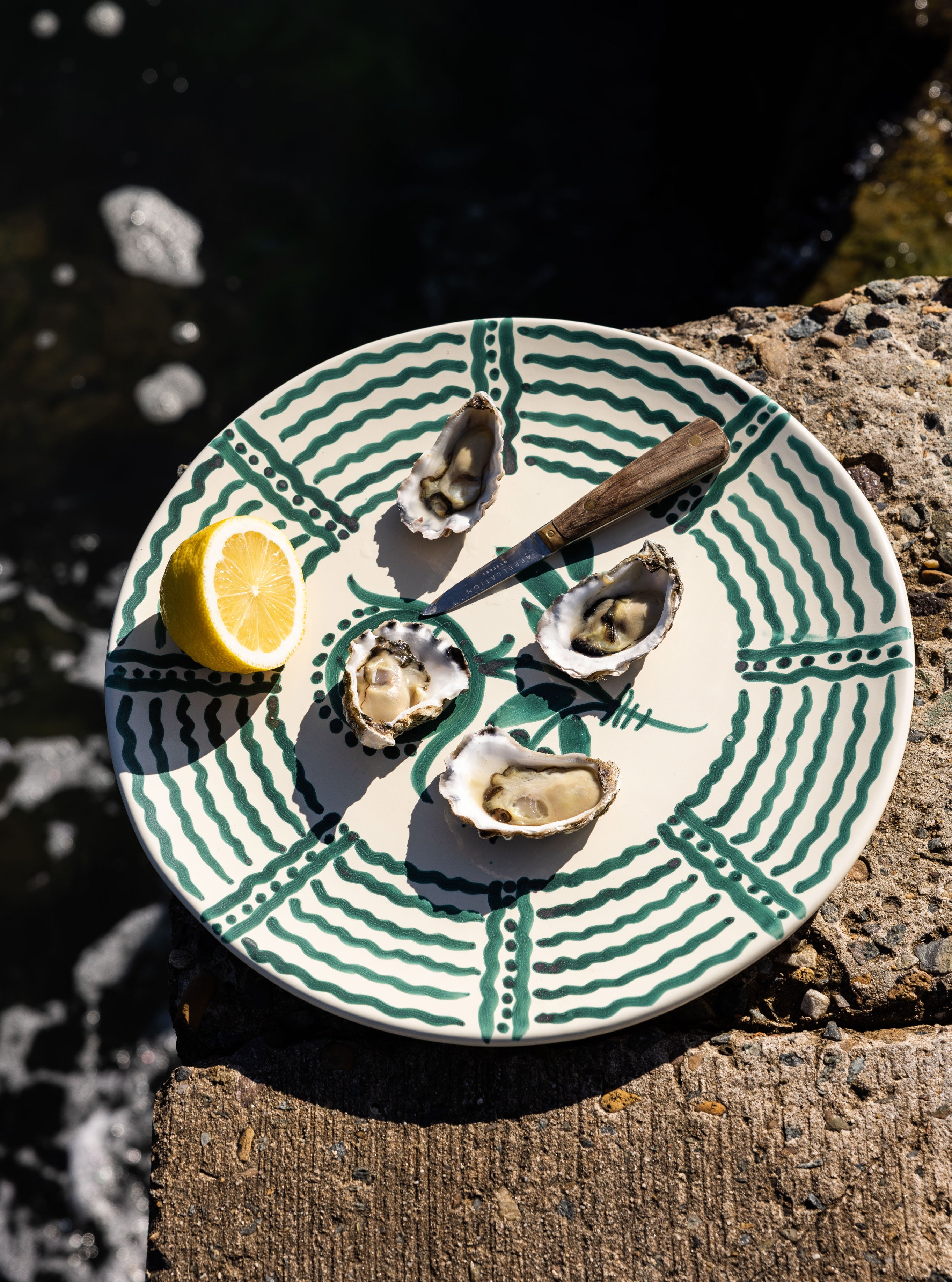 Four freshly shucked oysters with half a lemon and a shucking knife on a green patterned ceramic plate at the edge of a rock pool