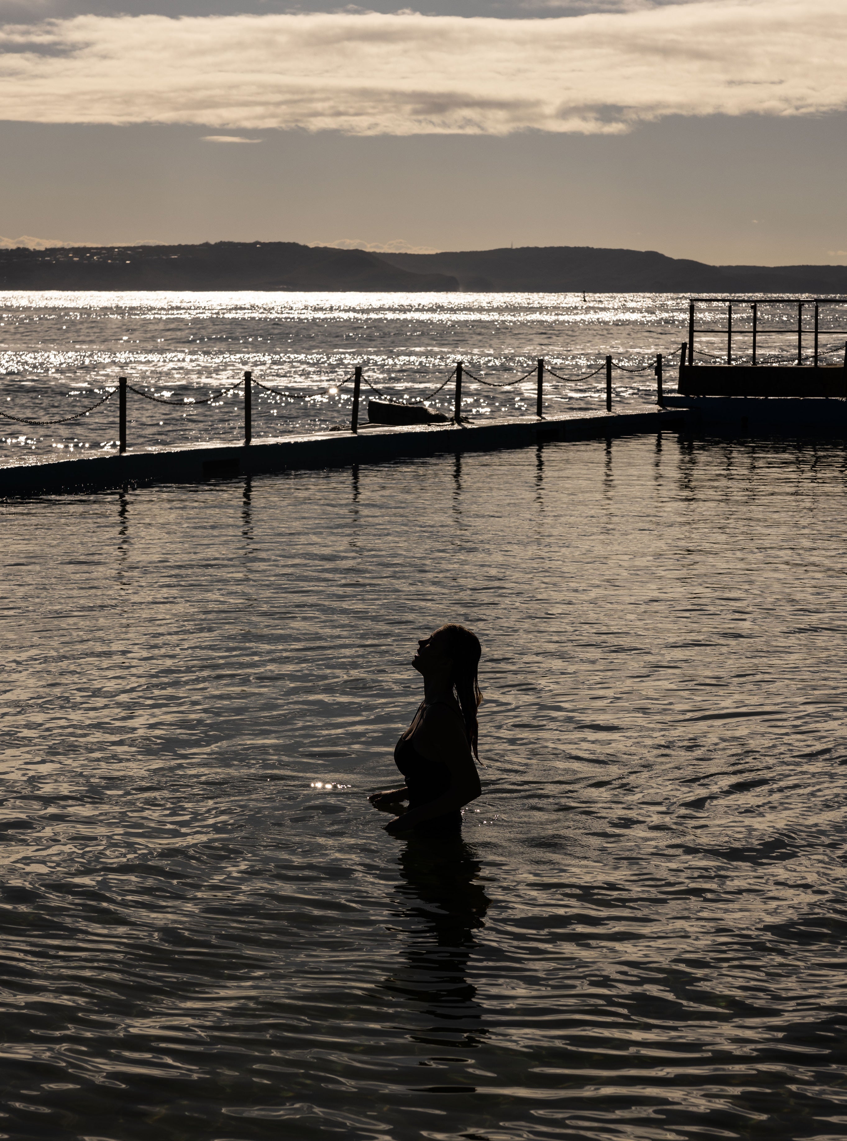 Silhouette of a woman standing waist-deep in a tidal ocean pool with sparkling backlit water and a headland in the distance