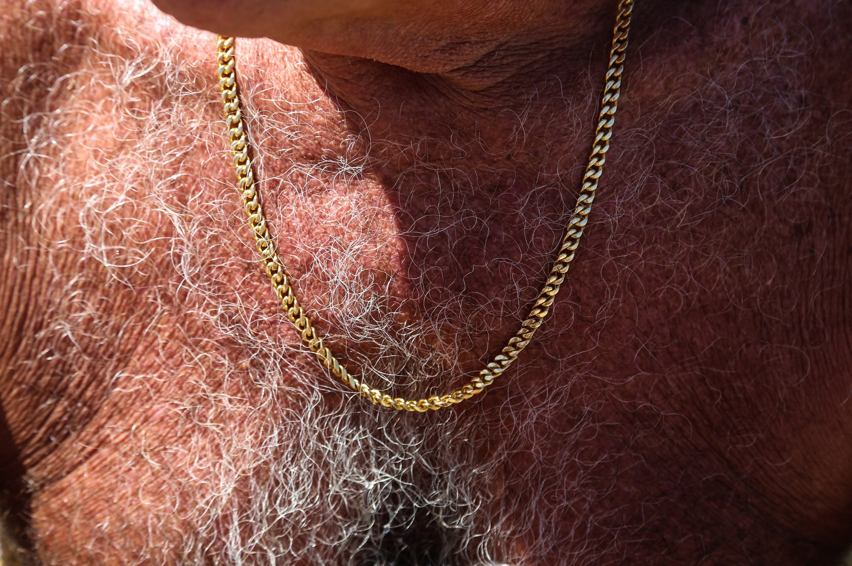 Extreme close-up of a gold curb chain necklace resting on a sun-tanned and weathered chest with white hair, evoking Bondi beach culture
