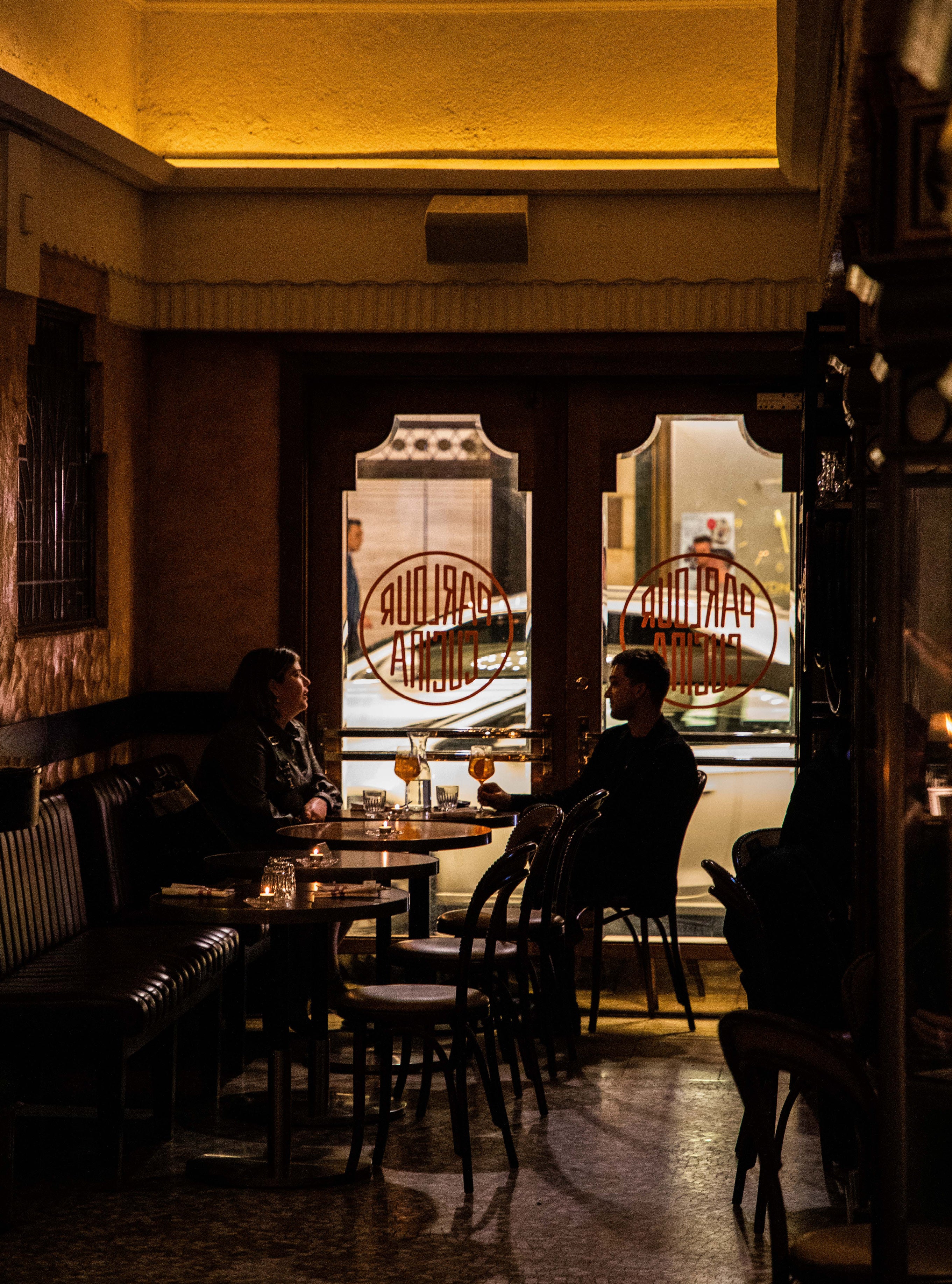 Moody interior of a vintage Sydney bar with two patrons seated at a table silhouetted against arched windows with reversed parlour signage