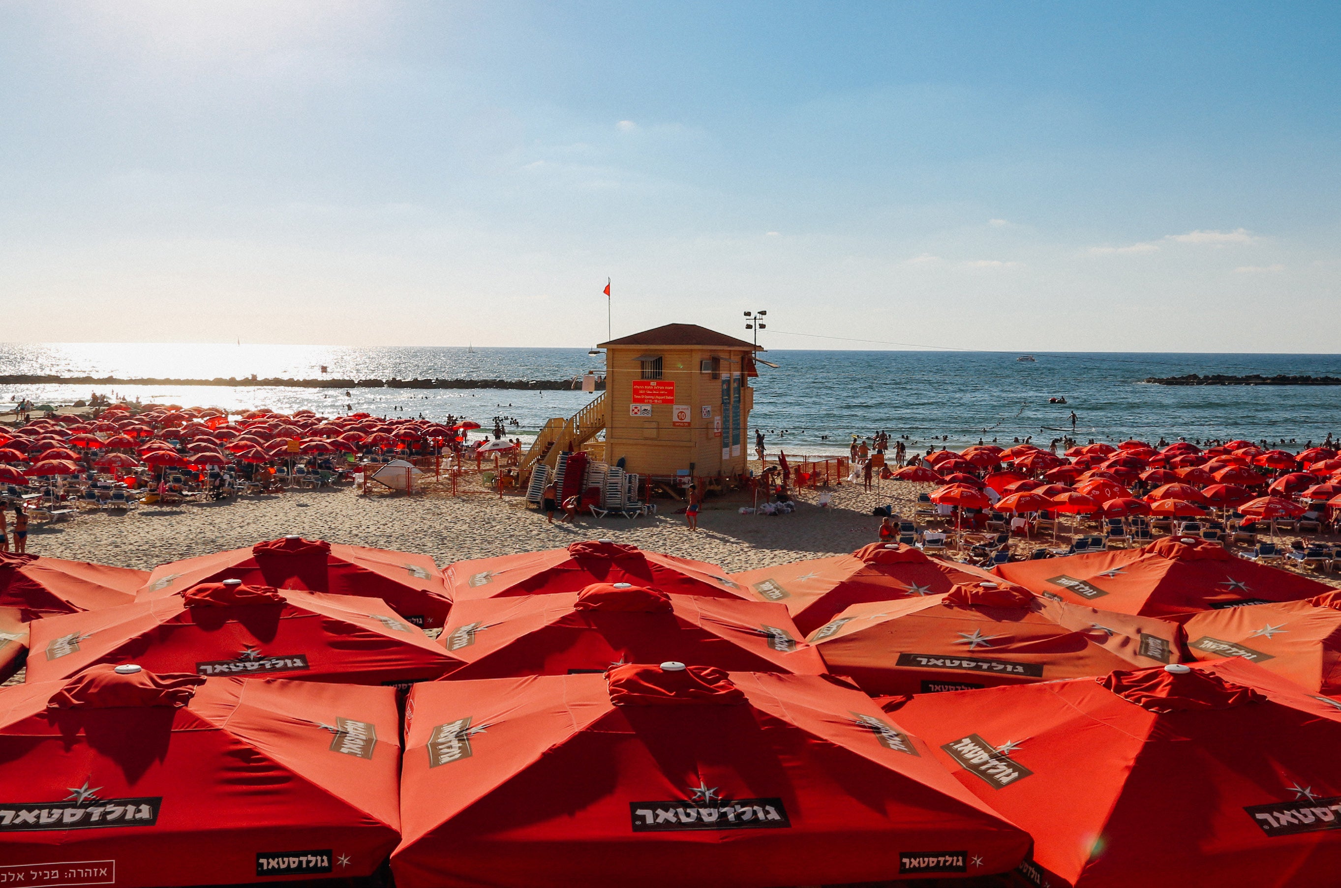 Dozens of red branded beach umbrellas filling the sand in front of a yellow lifeguard station on Gordon Beach Tel Aviv
