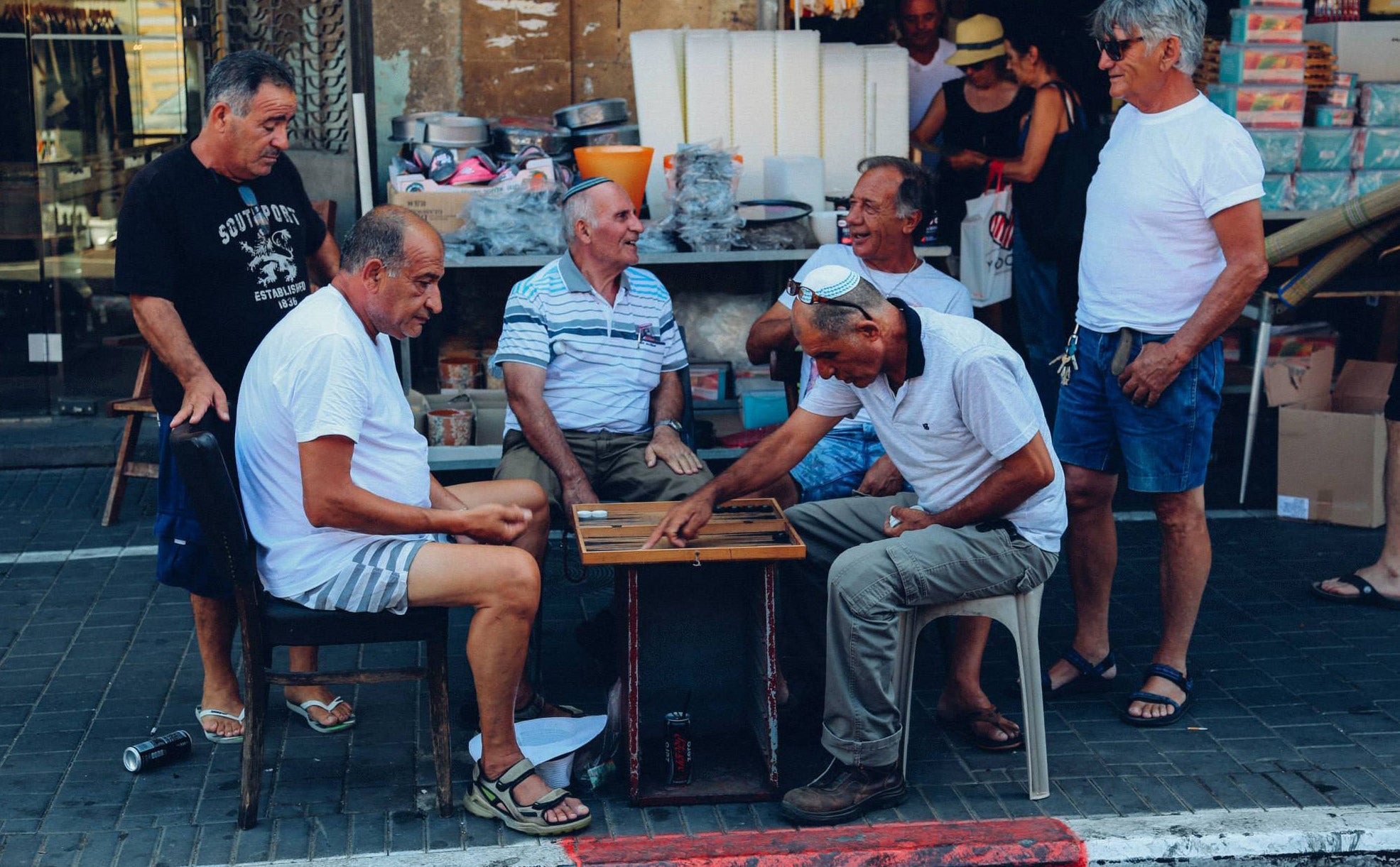 Group of elderly men sitting around a backgammon board on plastic chairs outside a market stall in Tel Aviv laughing and socialising