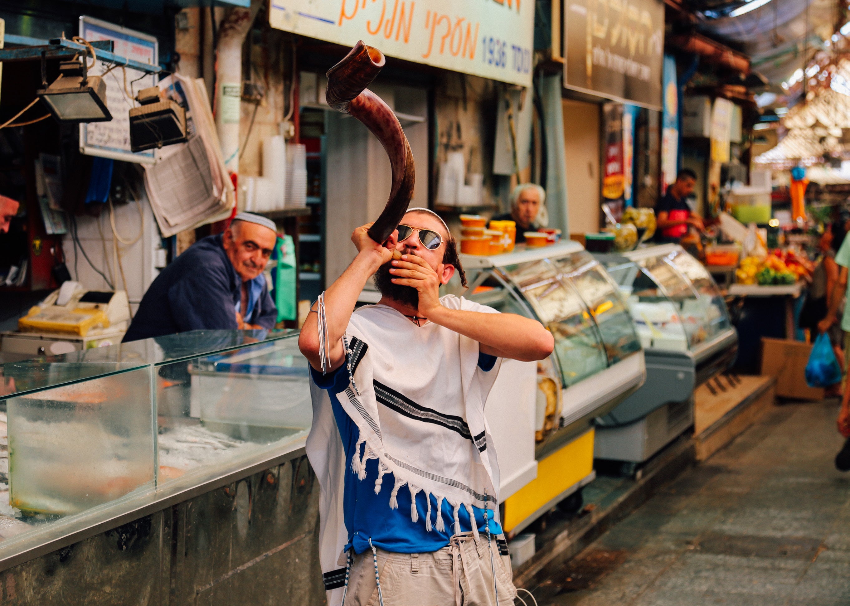 Young man wearing a tallit prayer shawl and sunglasses blowing a large curved shofar horn inside the Carmel Market with vendors watching