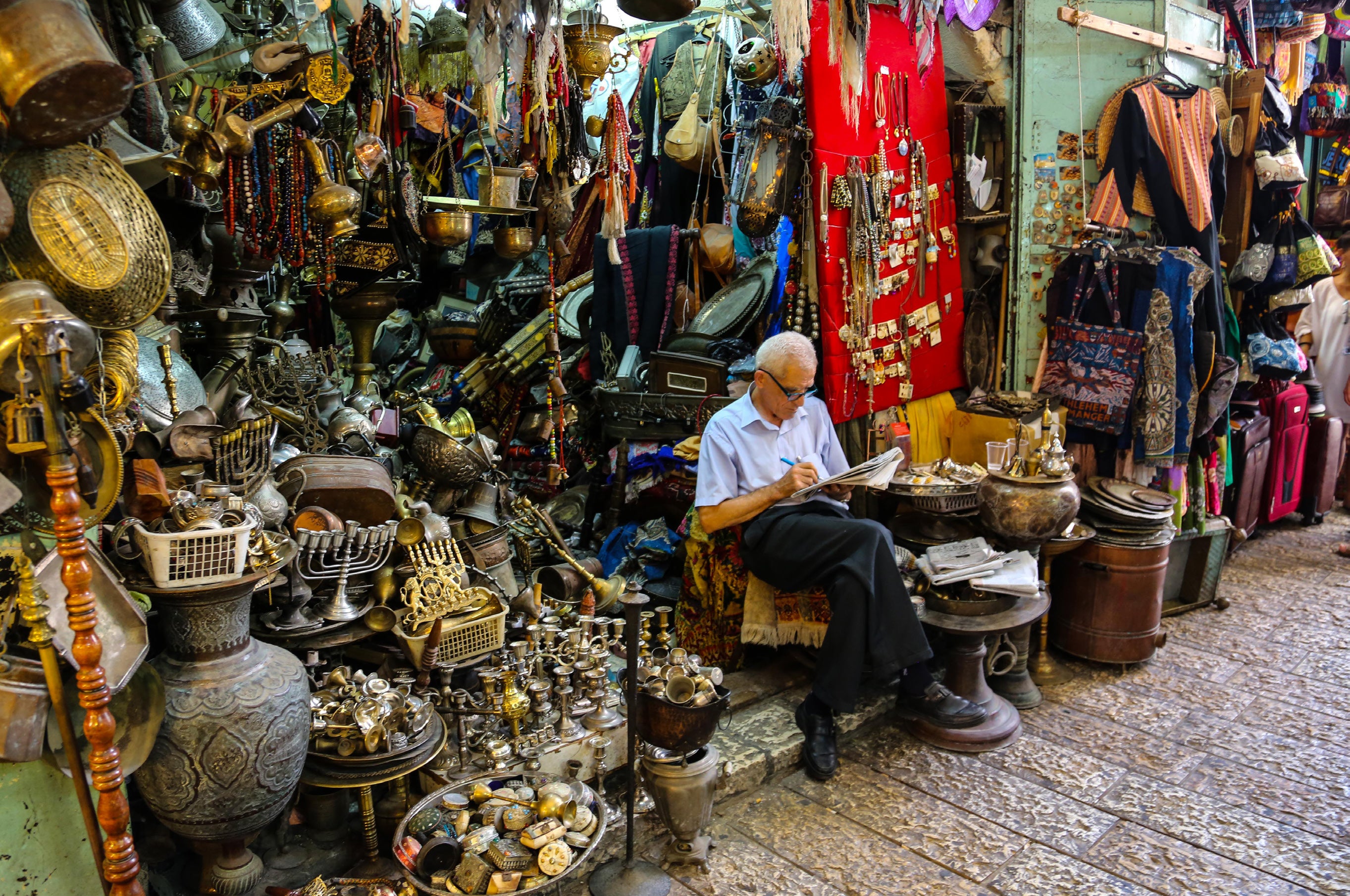 Elderly man sitting and reading surrounded by an enormous collection of brass antiques menorahs and curiosities at the Jaffa flea market