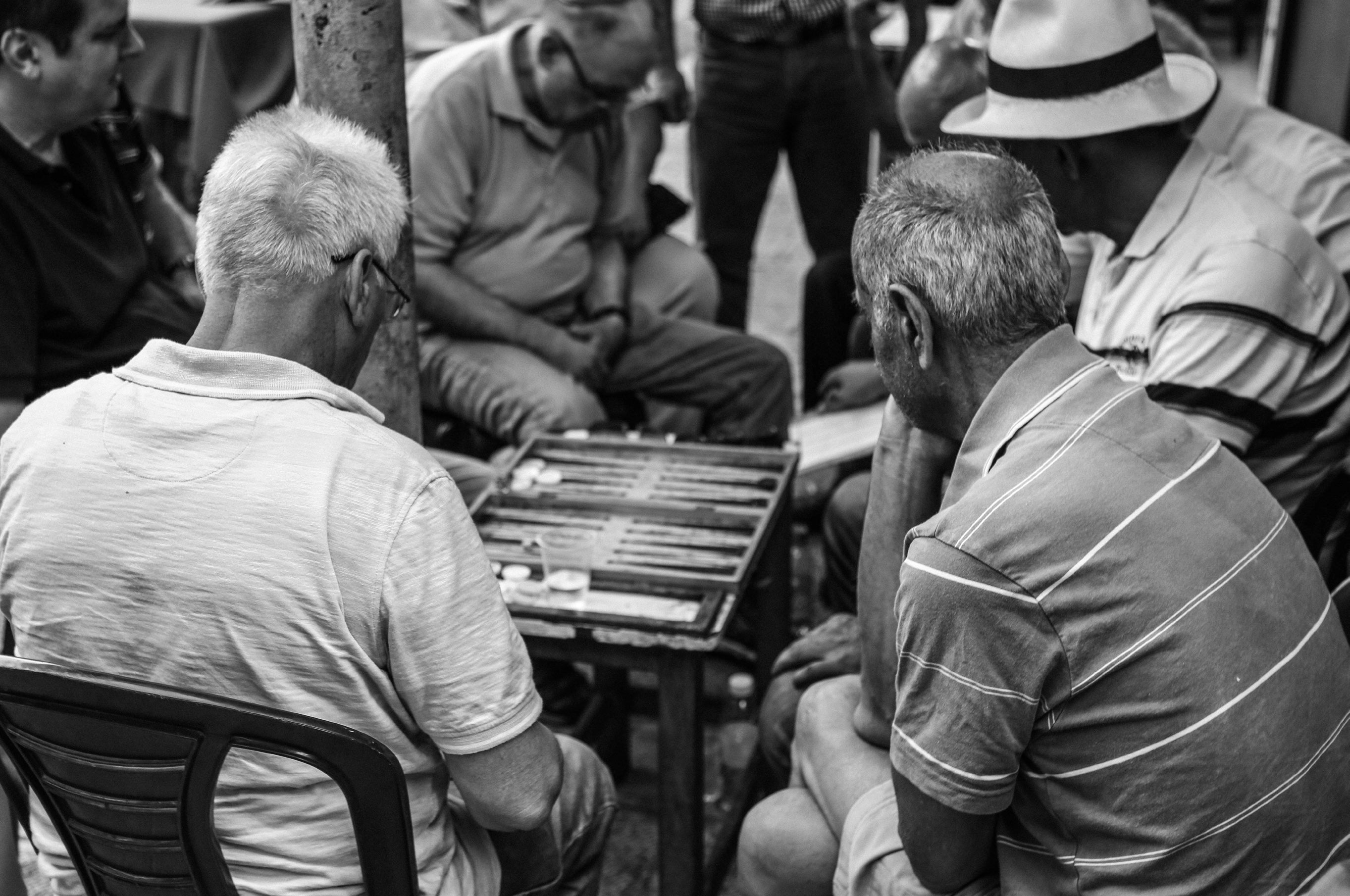 Black and white close-up of elderly men playing backgammon viewed from behind their shoulders showing the board between them
