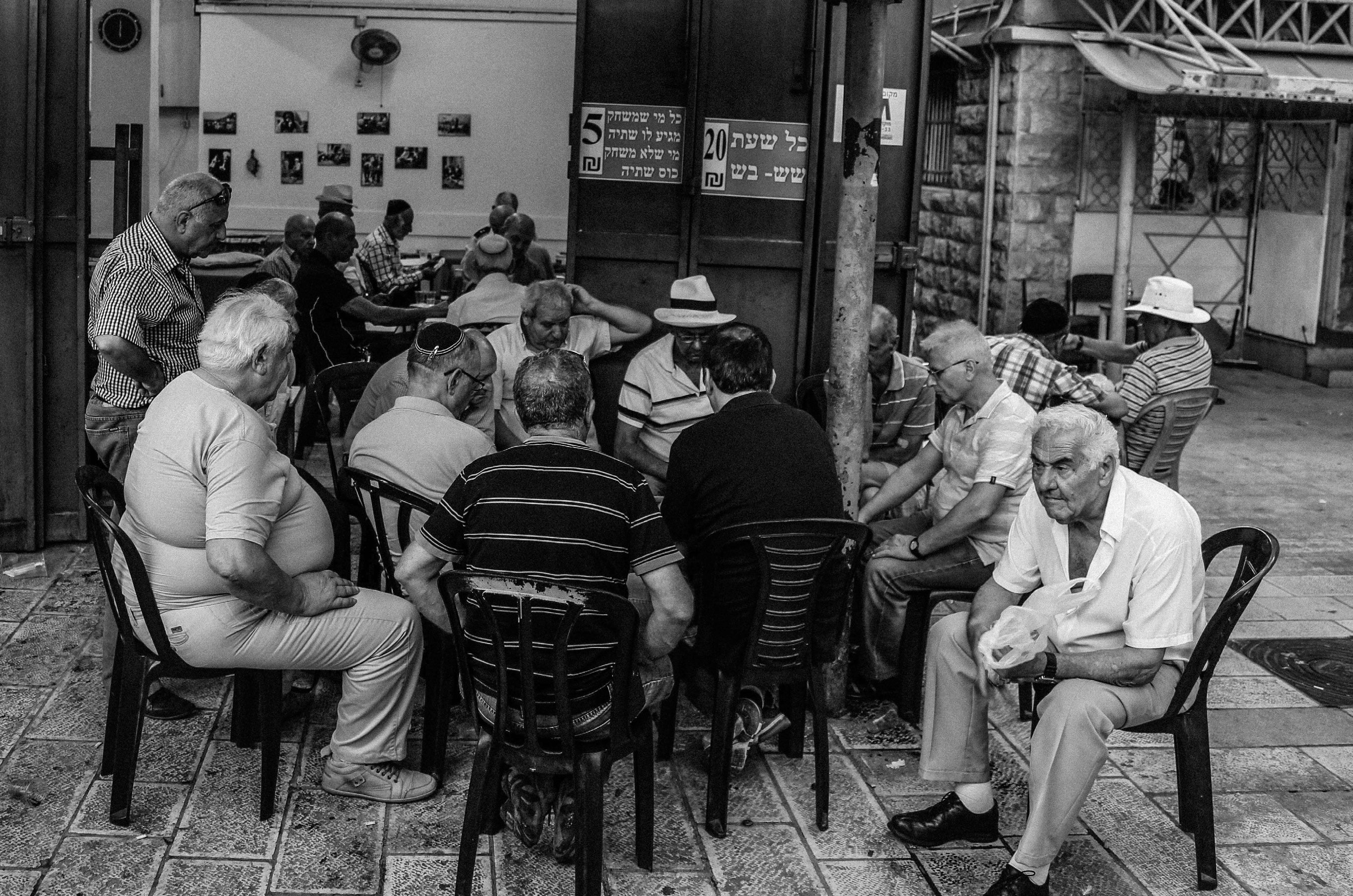 Black and white photograph of a large group of elderly men sitting in a circle on plastic chairs at an outdoor courtyard gathering in Tel Aviv
