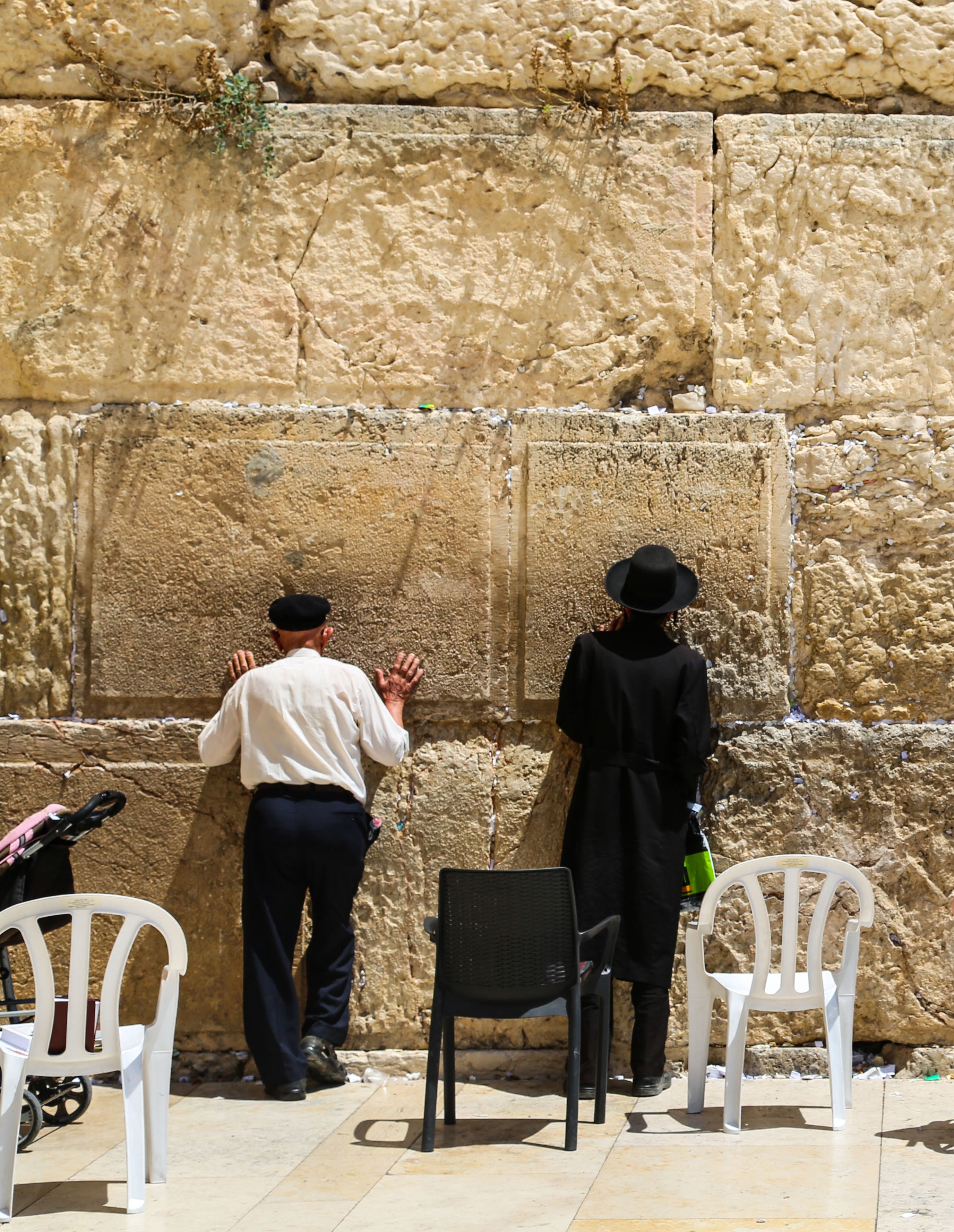 Two men in dark hats praying with hands touching the ancient limestone blocks of the Western Wall in Jerusalem with empty plastic chairs nearby