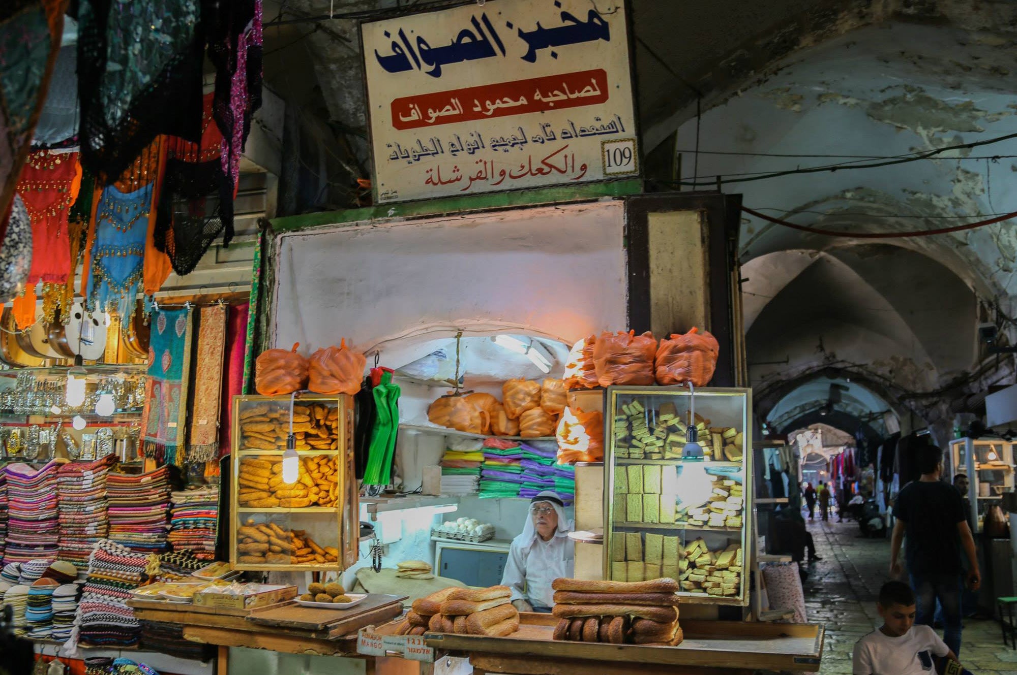 Young baker sitting behind a counter displaying fresh bread and pastries under an Arabic bakery sign in the Old City of Jerusalem