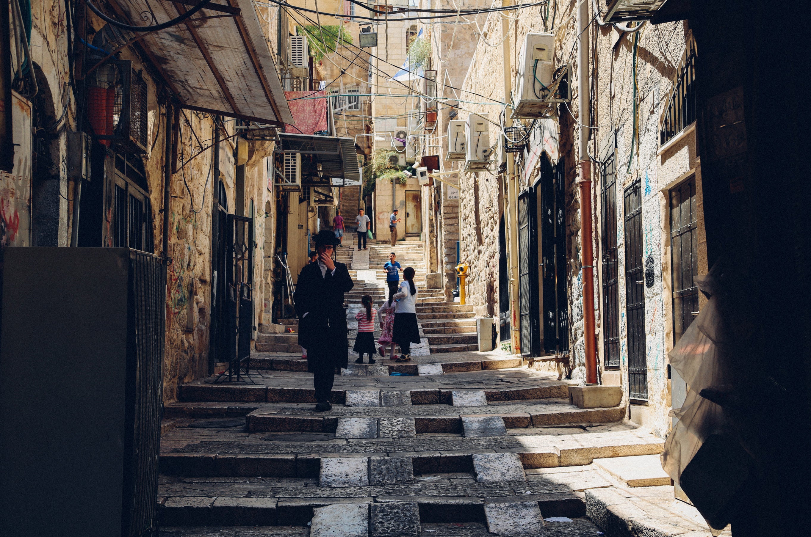 Orthodox man walking down a narrow stepped stone alleyway in the Old City of Jerusalem with a family visible further up the stairs