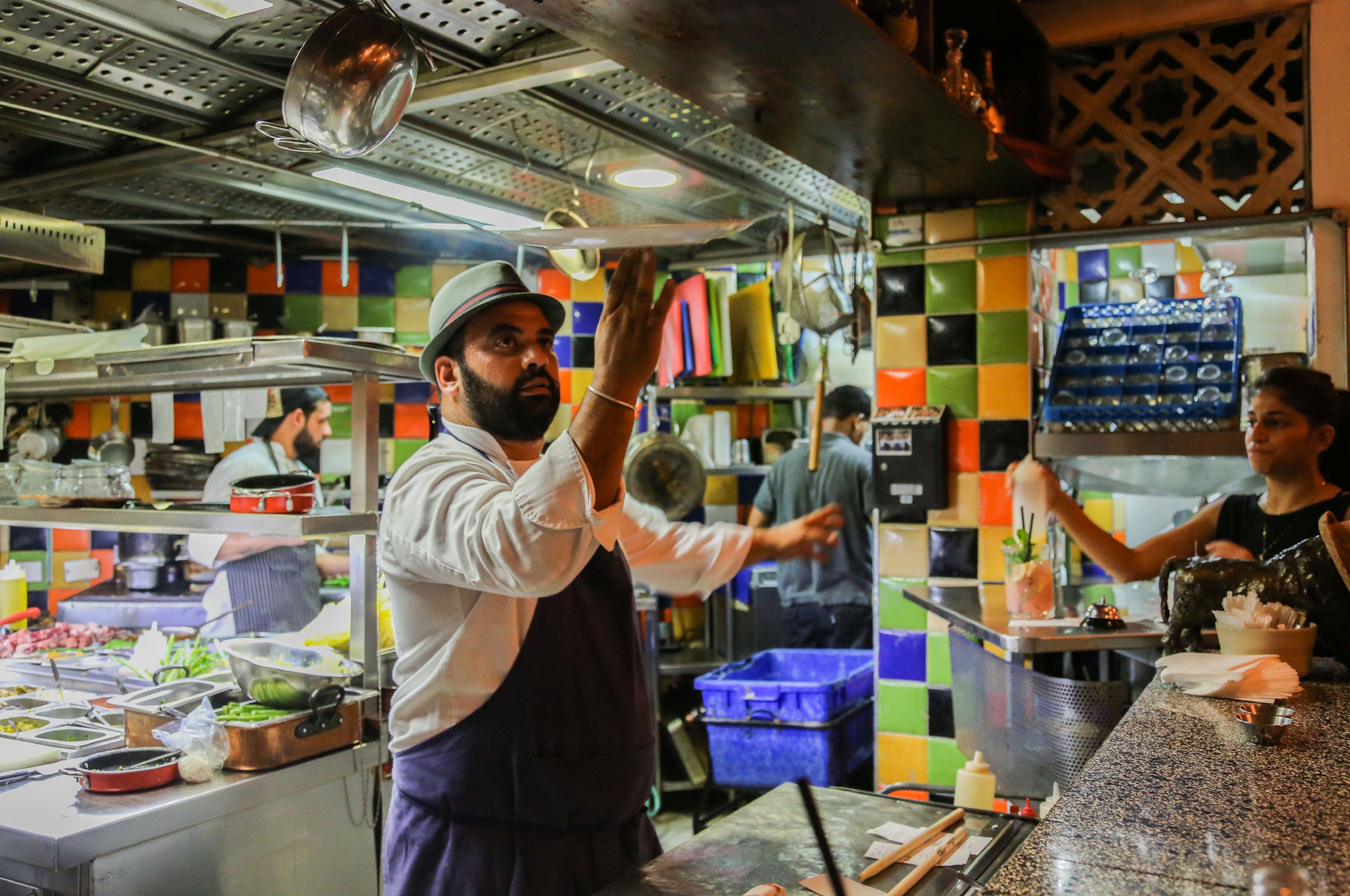 Chef reaching up to grab a pan from an overhead shelf in a bustling restaurant kitchen with colourful tiled walls in Tel Aviv
