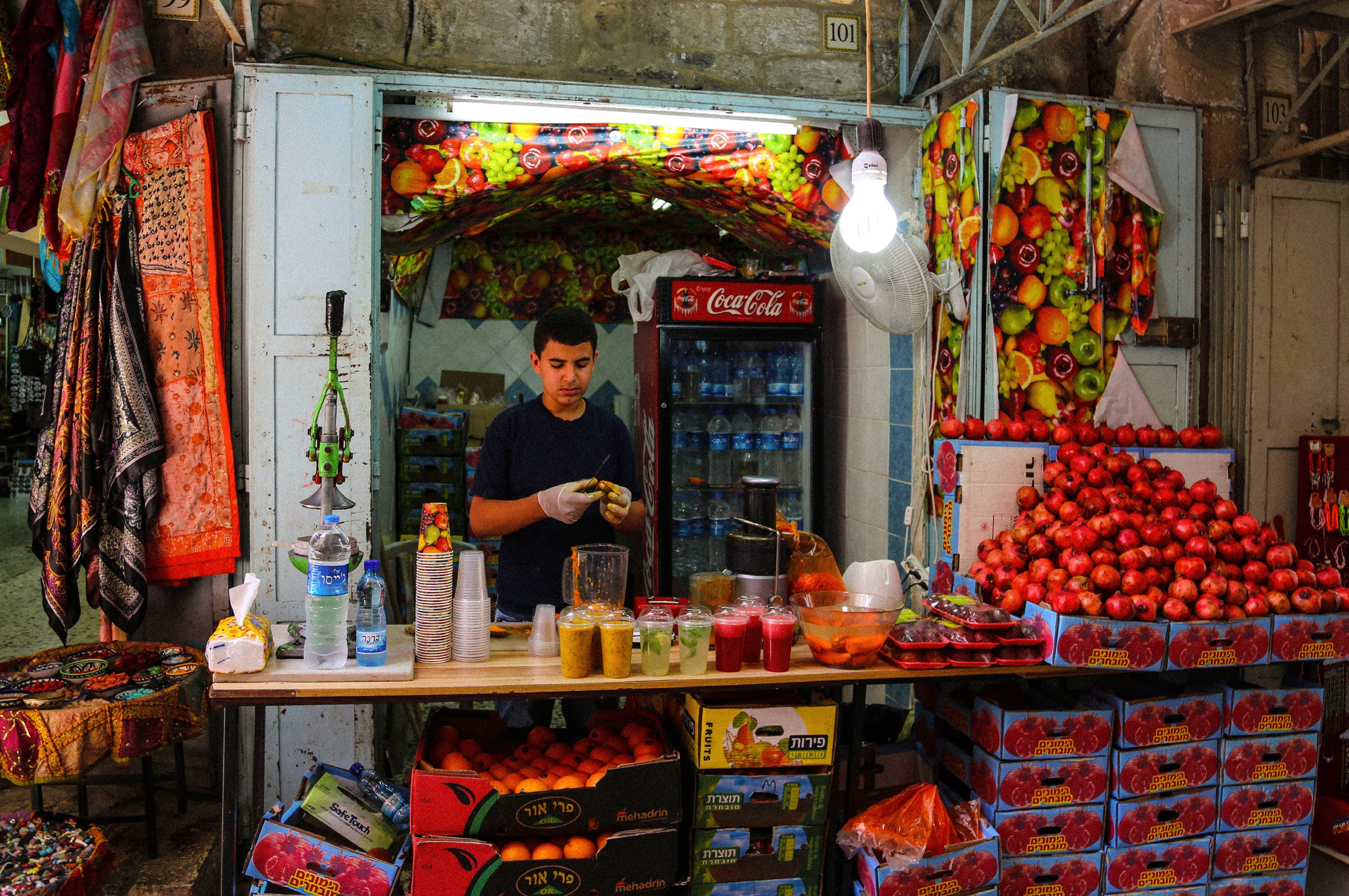 Young vendor preparing fresh juice at a small kiosk surrounded by stacked crates of pomegranates and oranges in the Old City market