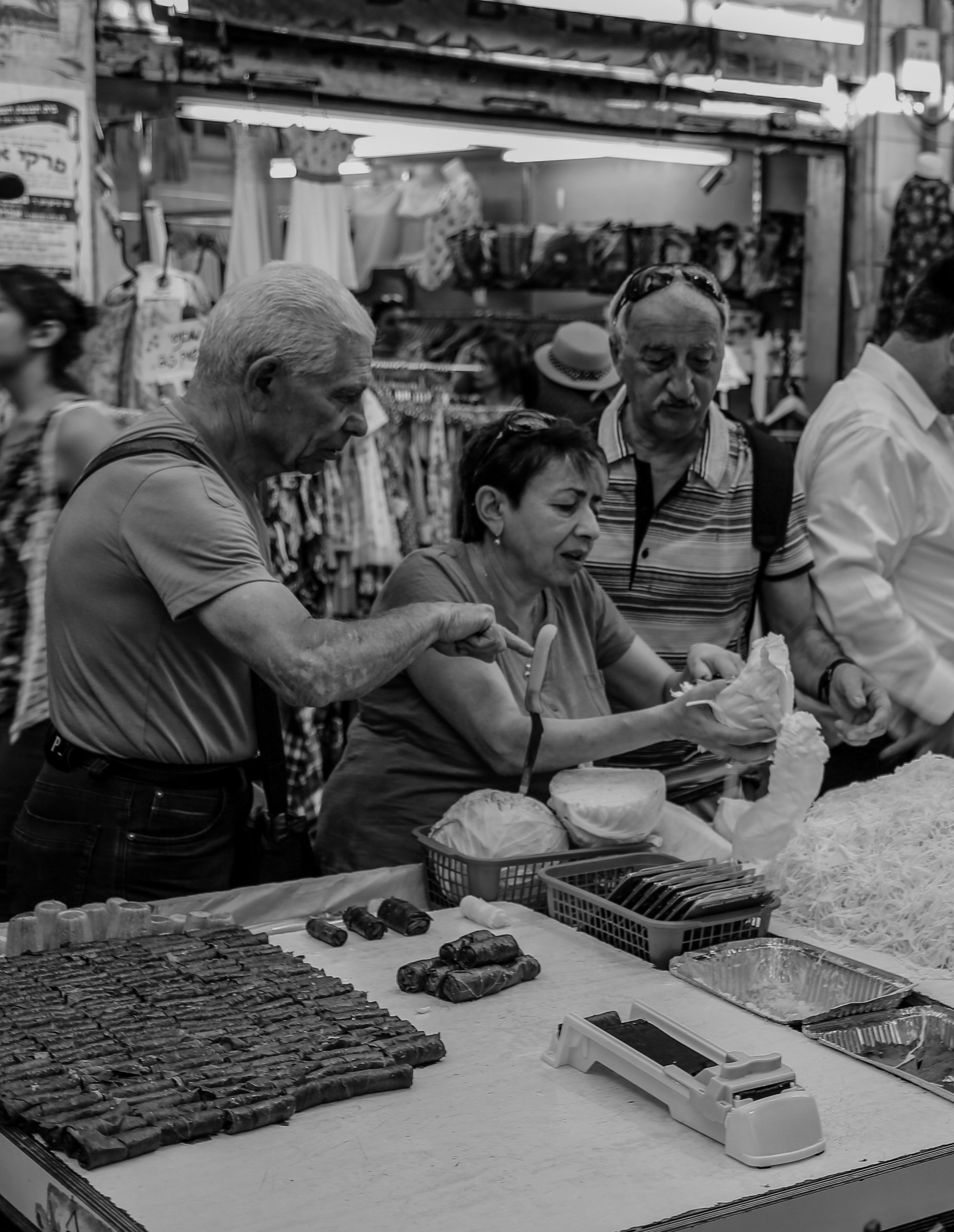 Black and white photograph of a vendor serving customers at a market stall counter displaying trays of baklava and Middle Eastern pastries