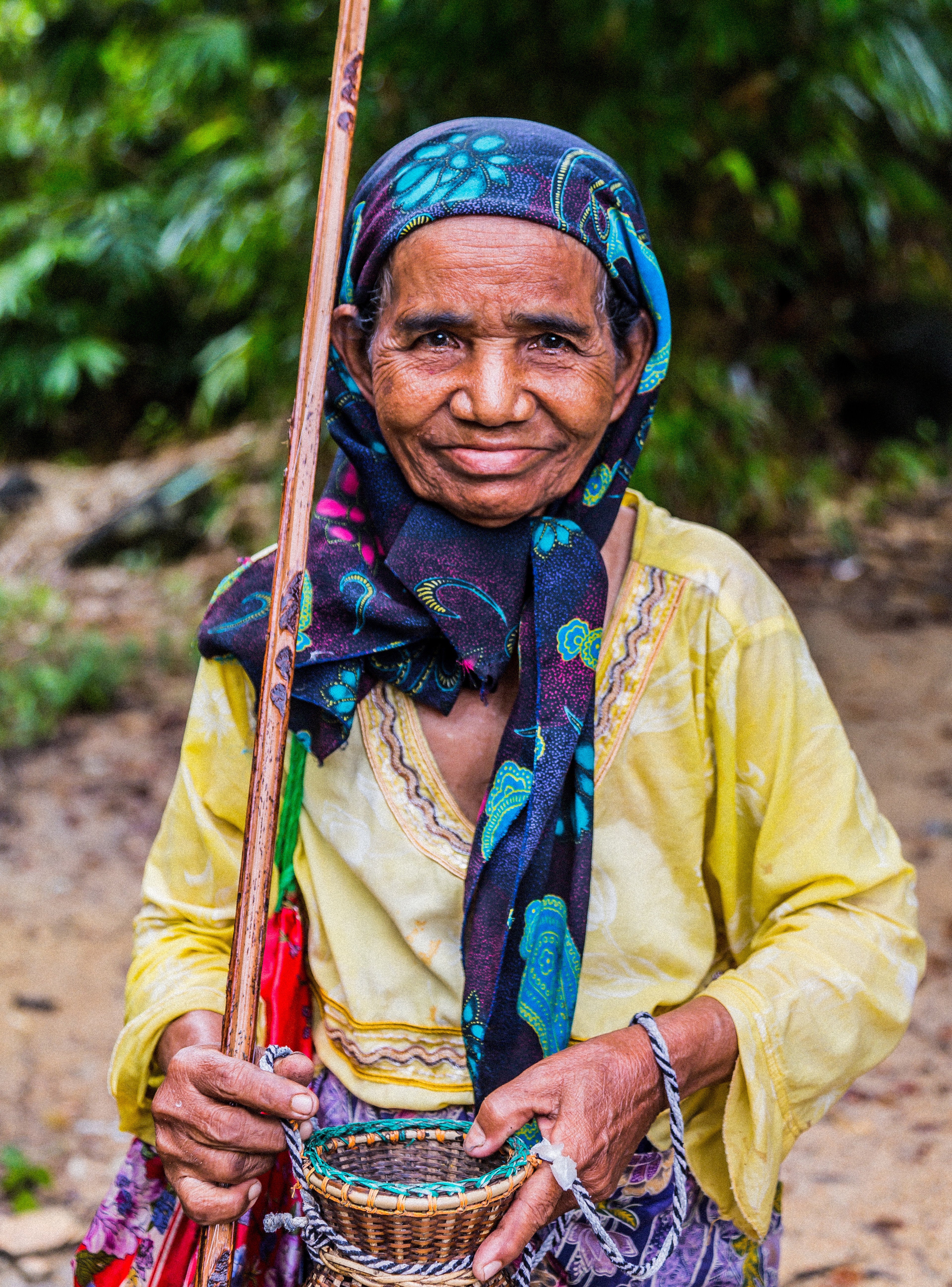 Elderly Thai woman in yellow shirt and blue patterned headscarf holding a wooden walking stick and small woven basket in lush green jungle