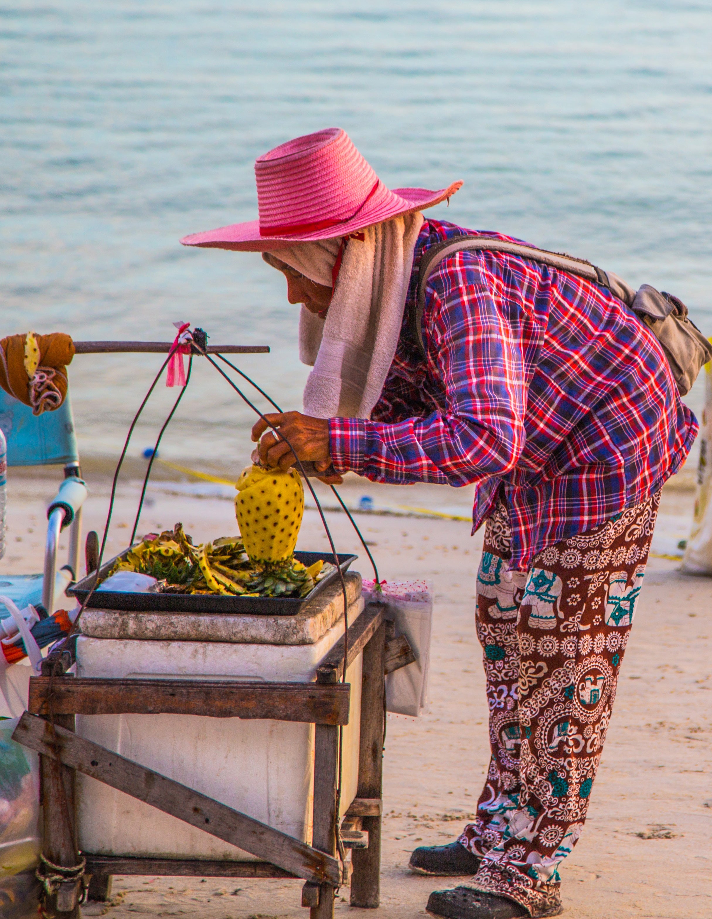 Thai woman in pink hat and colourful plaid shirt arranging pineapple and fruit on a portable cart on a sandy beach with ocean behind