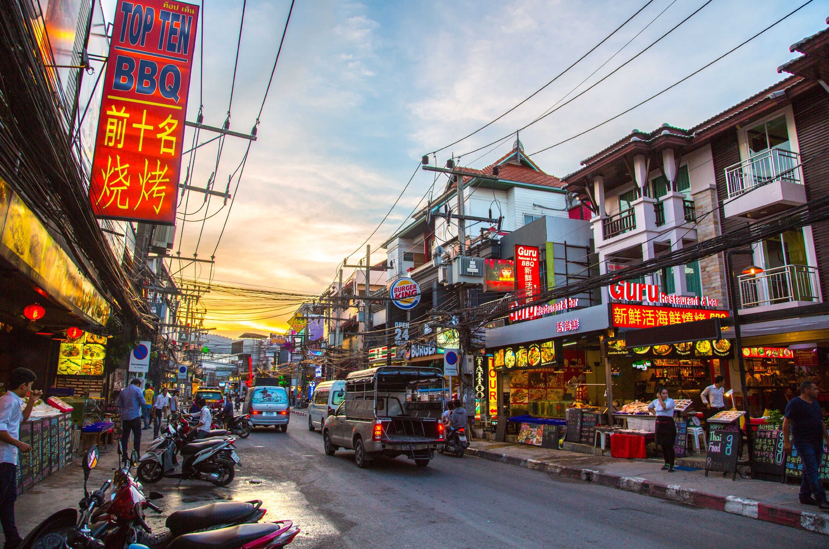 Bustling Thai town street at sunset lined with neon signs in Thai and Chinese script, parked motorbikes, power lines and a warm golden sky