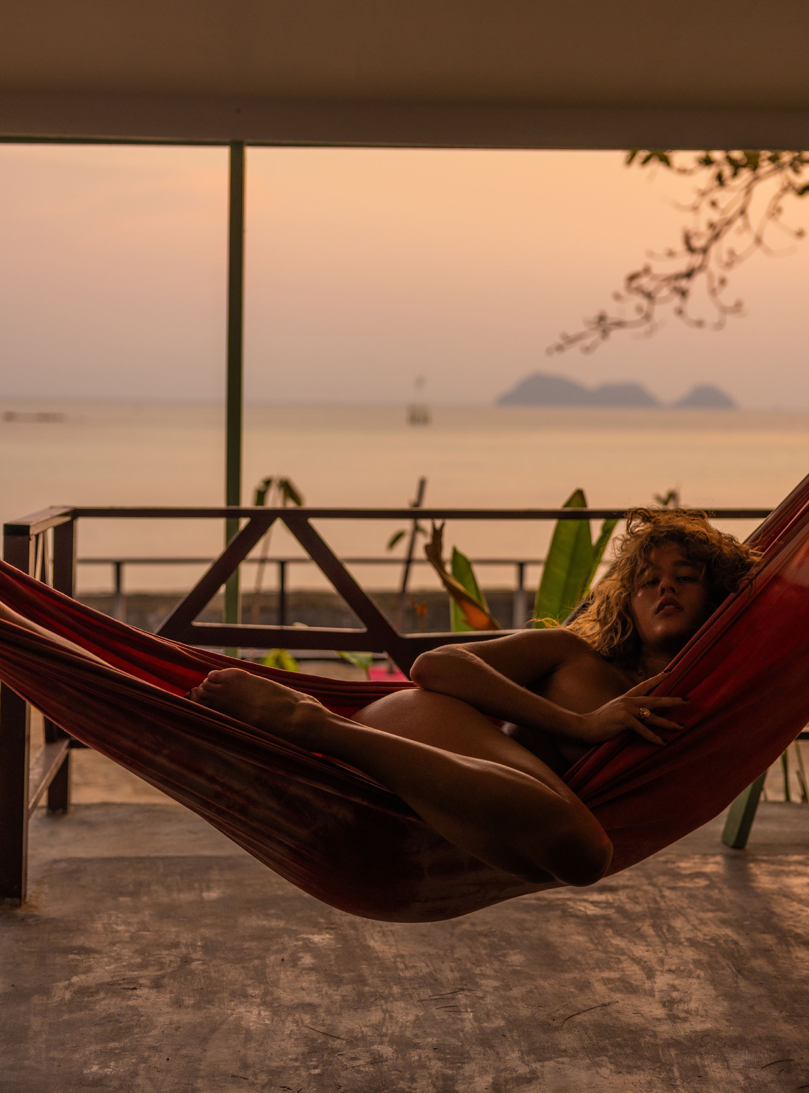 Woman reclining in a red striped hammock on a covered balcony with a view of distant islands and calm ocean at pink dusk