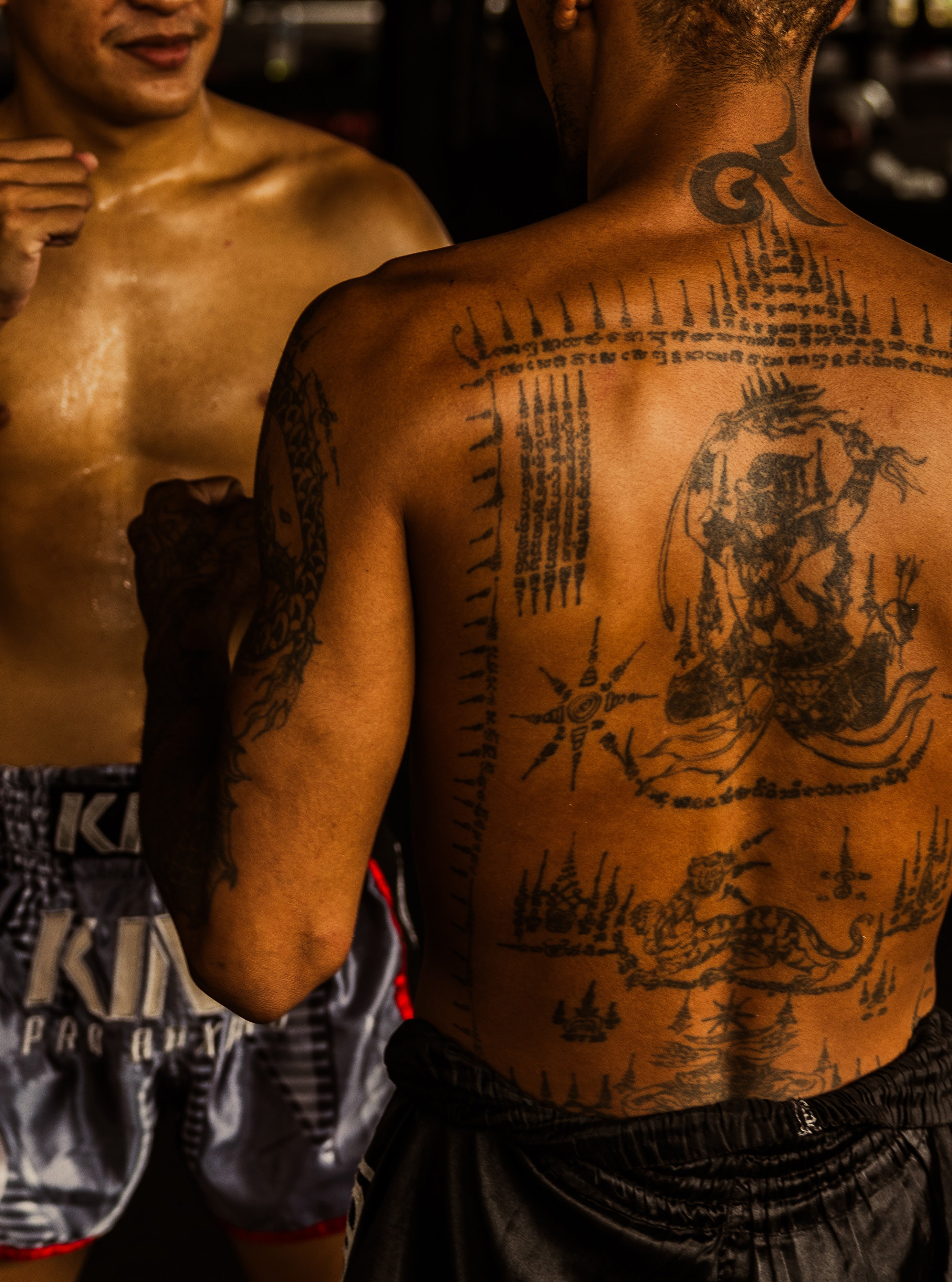 Close-up of a Muay Thai fighter's sweaty back covered in intricate Sak Yant sacred tattoos with another fighter's torso visible beside him