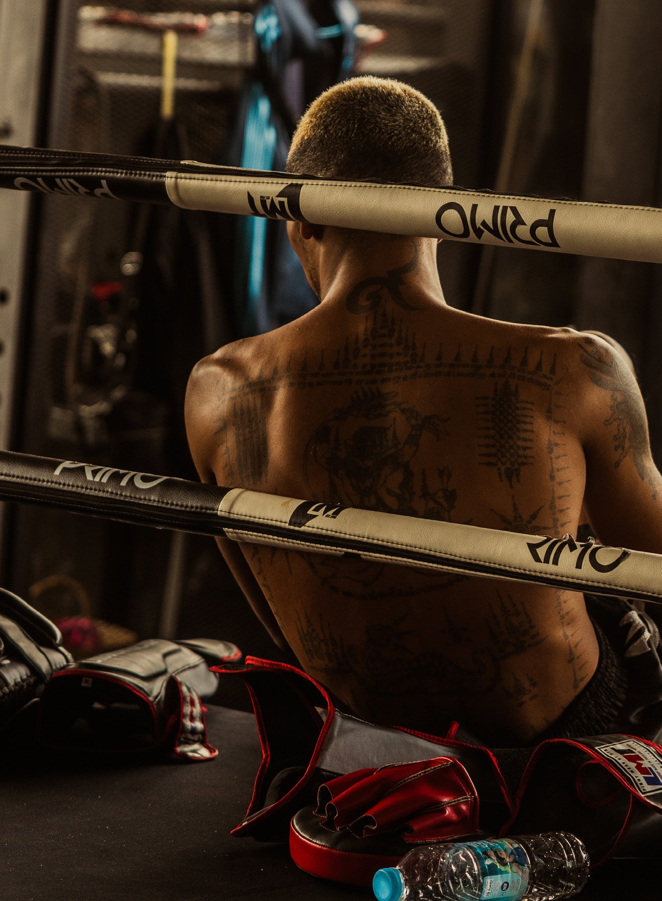 Rear view of a tattooed Muay Thai fighter with bleached hair sitting in the corner of a ring leaning on the ropes with gloves and gear beside him