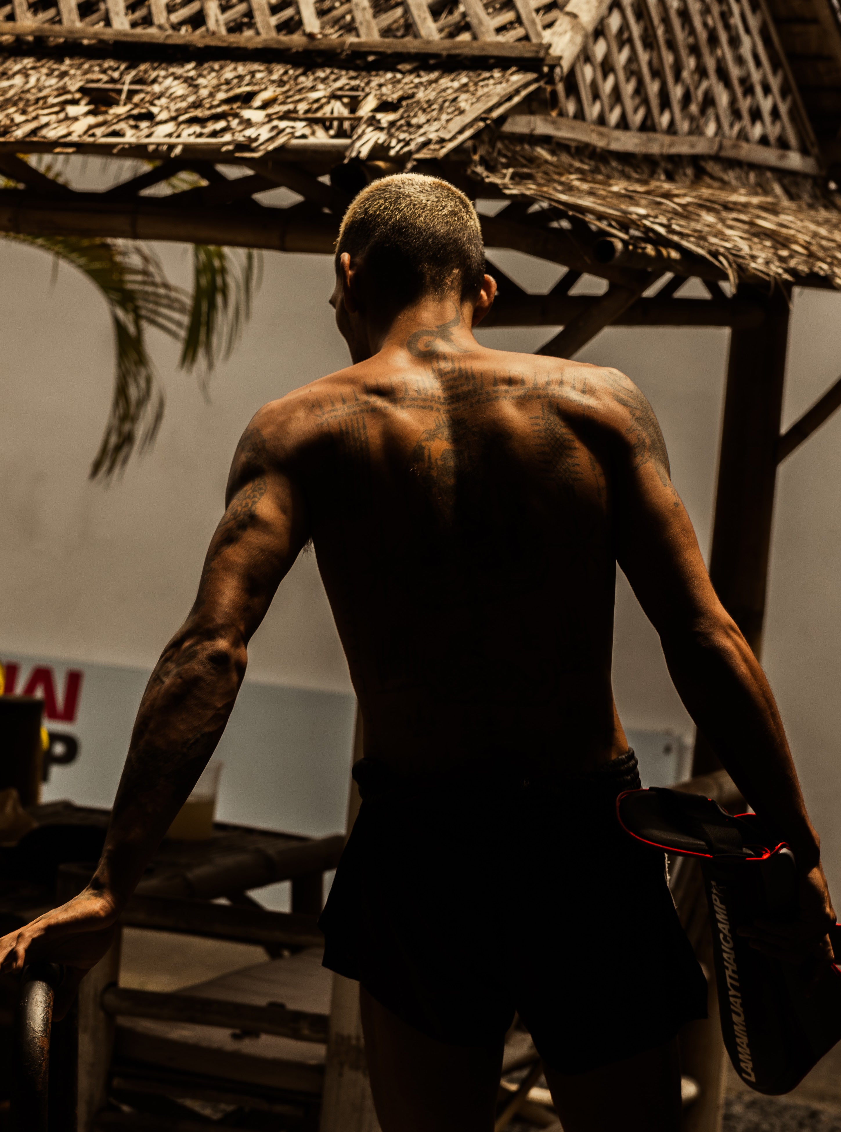 Rear view of a muscular Muay Thai fighter with bleached hair and tattooed back walking away from camera under a thatched roof lit by warm light