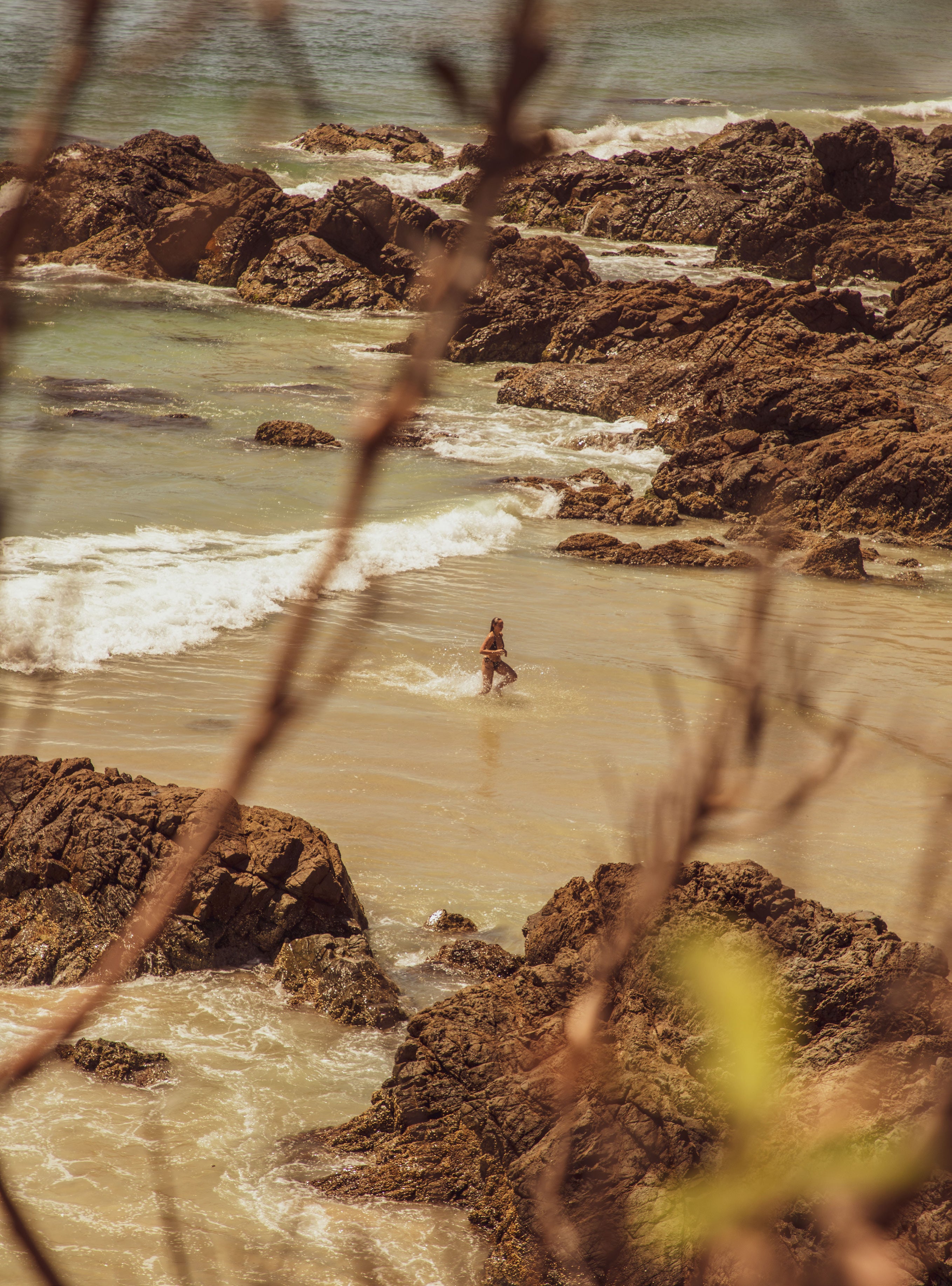 Warm-toned view through branches of a lone swimmer wading in a sandy cove surrounded by large coastal rocks