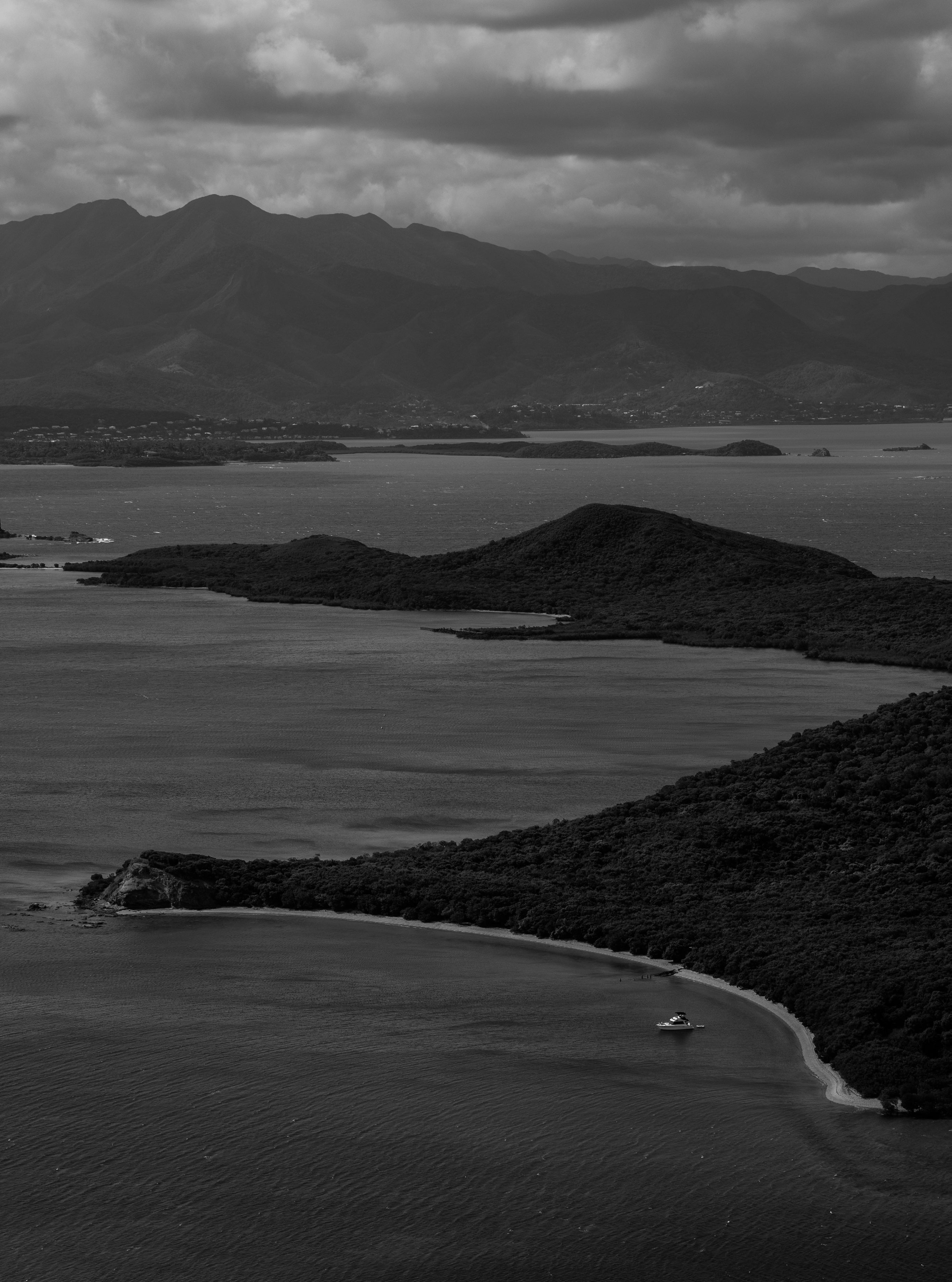 Black and white elevated view of a tropical archipelago with forested islands, calm dark water, a single boat and distant mountains under dramatic clouds