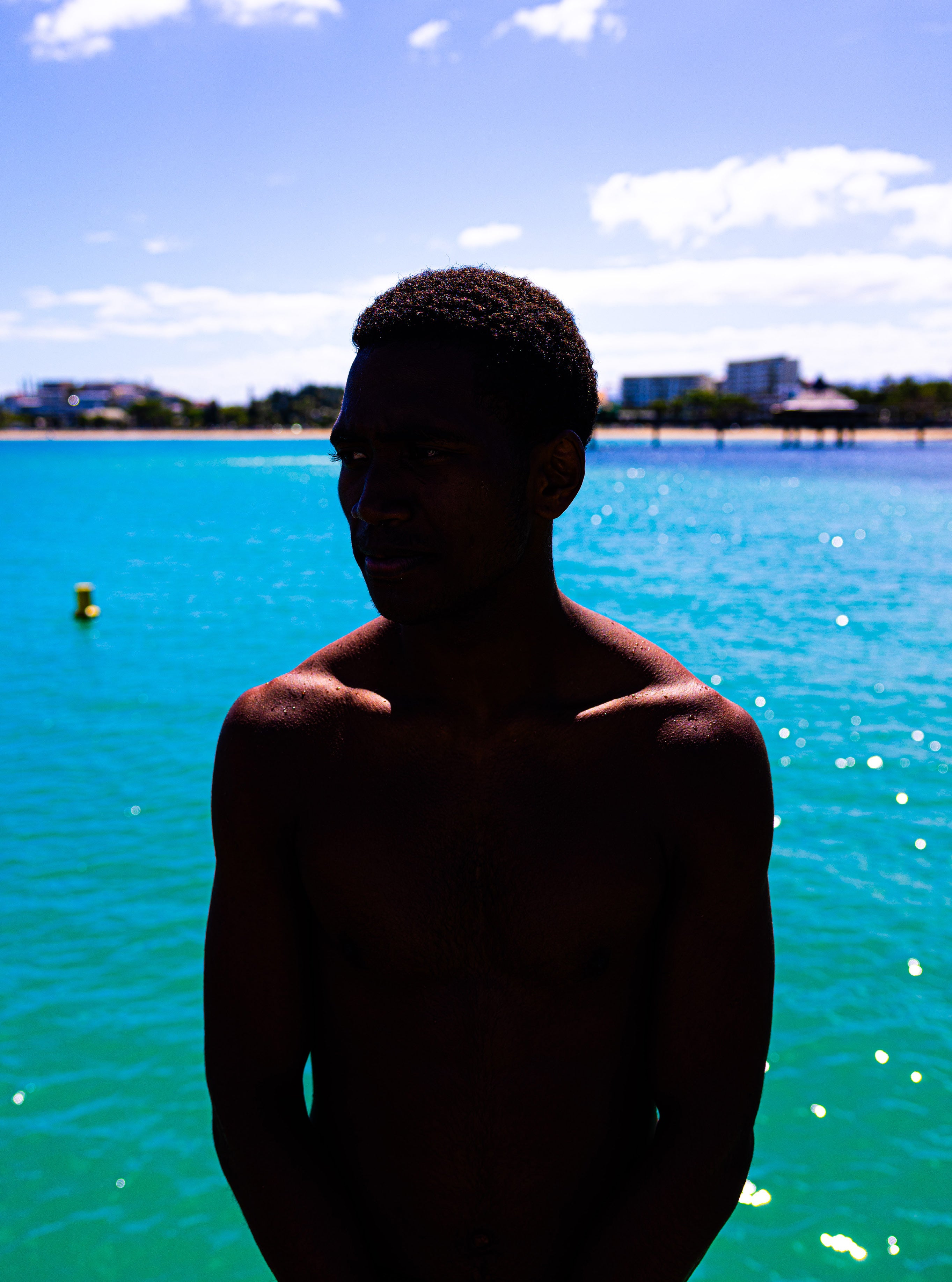 Silhouetted portrait of a young man standing in front of bright turquoise ocean water under a blue sky with scattered clouds