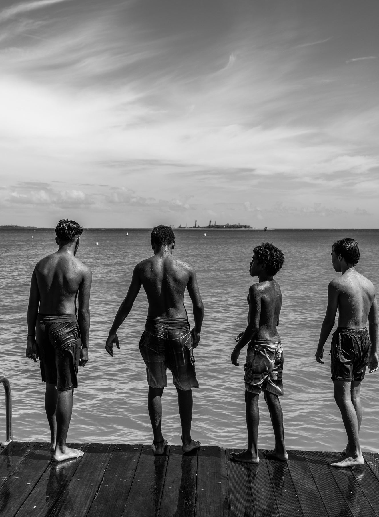 Black and white image of four boys standing at the edge of a wooden wharf looking out over calm water with a ship on the horizon