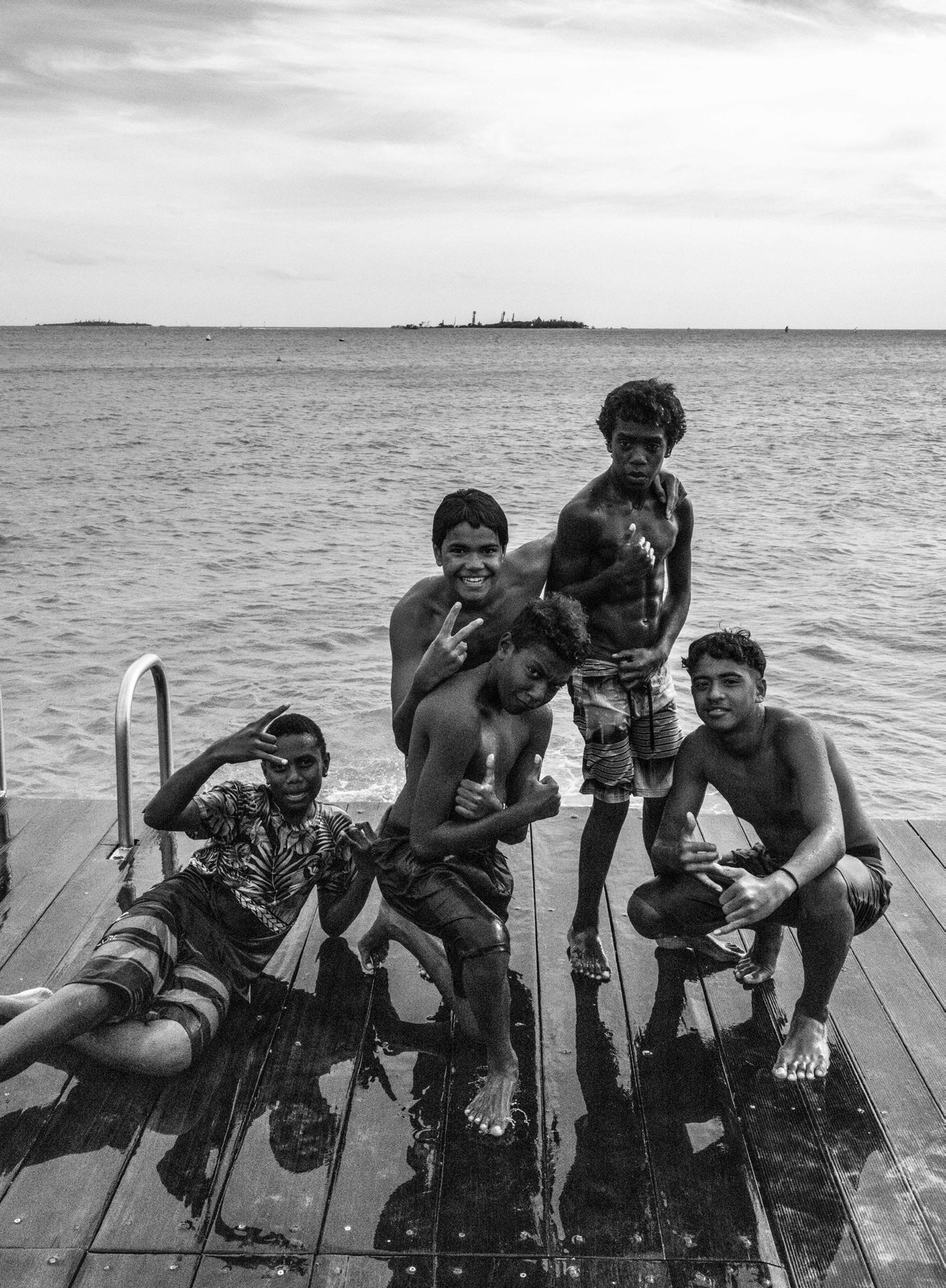 Black and white group portrait of six smiling island boys posing together on a wet wooden wharf with ocean behind them