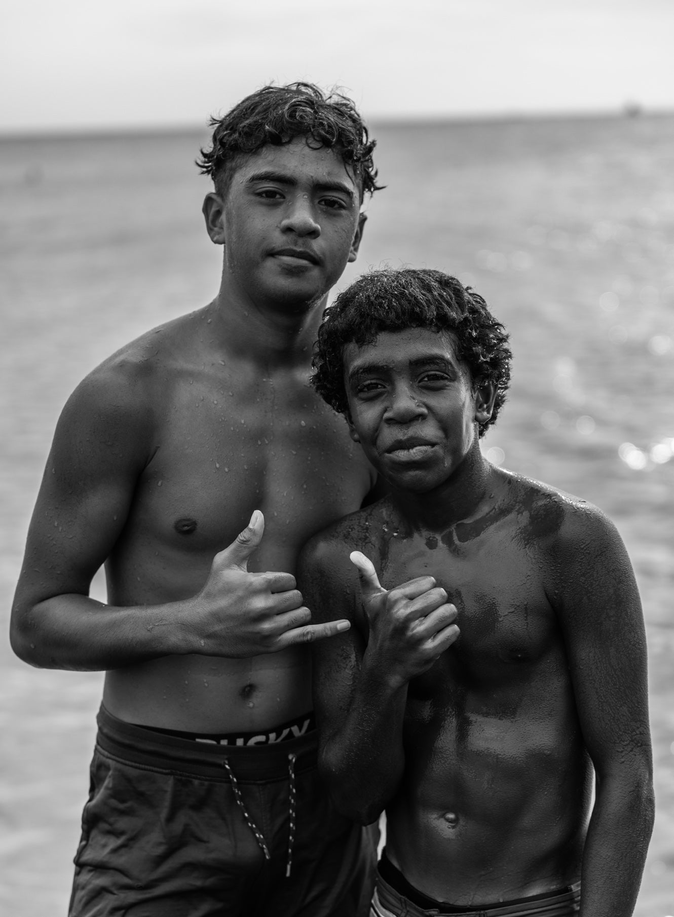 Black and white close portrait of two young island boys giving shaka hand signs with wet skin and the ocean sparkling behind them