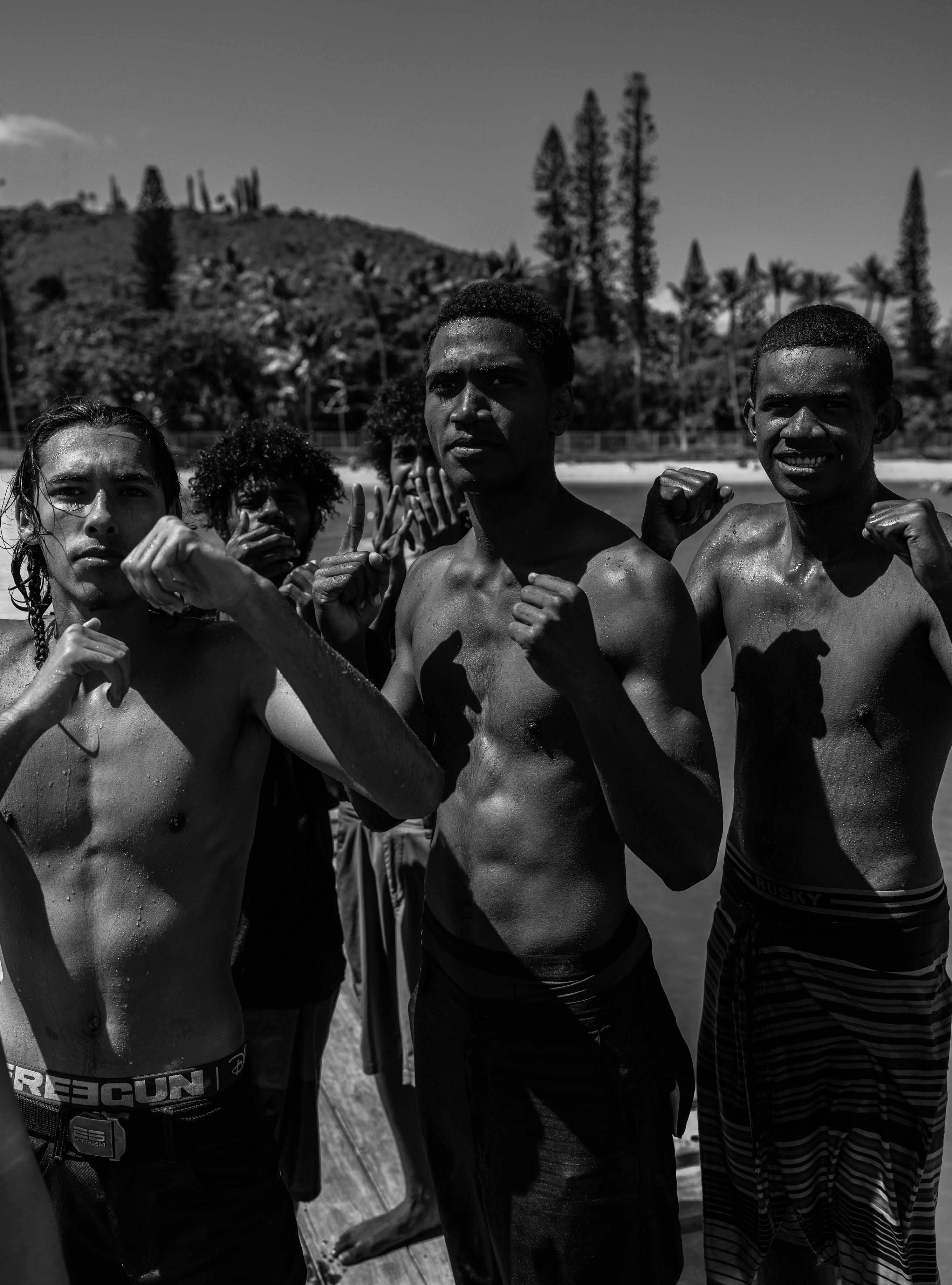 Black and white portrait of three young shirtless men posing with fists raised in a fighting stance with tall pine trees and hills behind