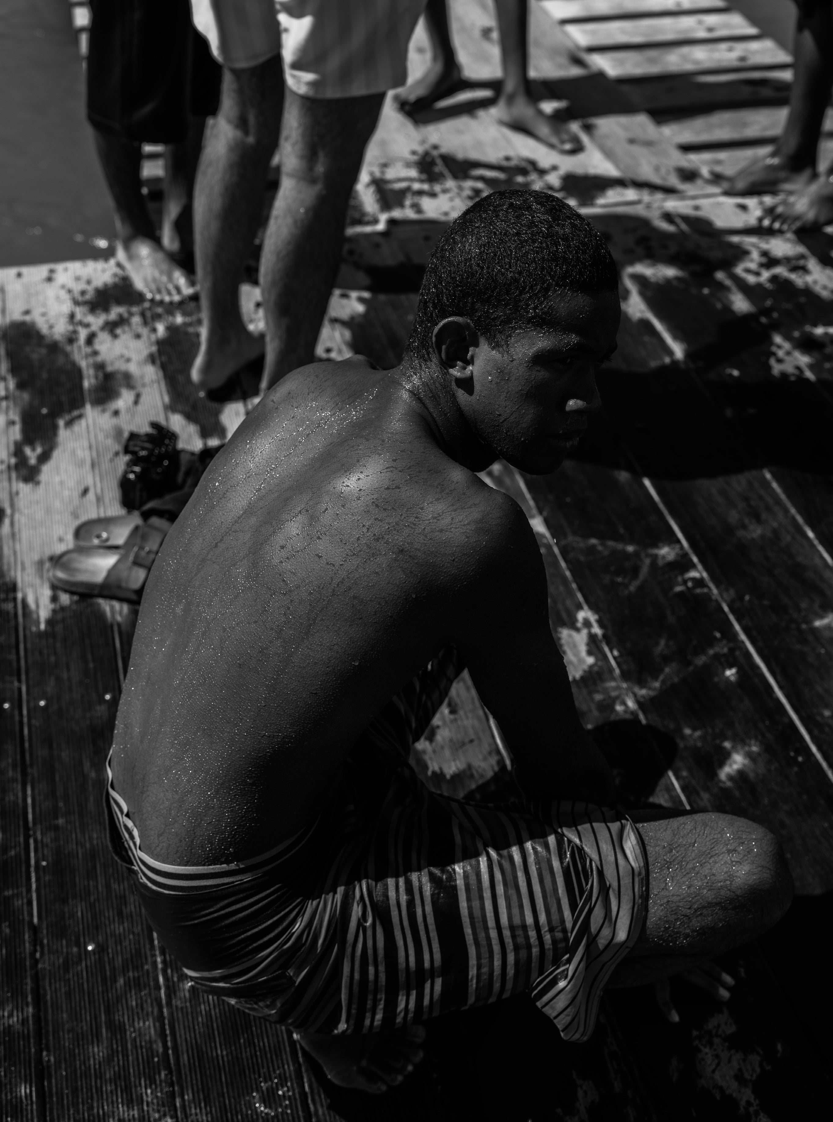 Black and white photograph of a boy sitting on a wet wooden dock with his back to the camera, wet skin glistening, people standing behind
