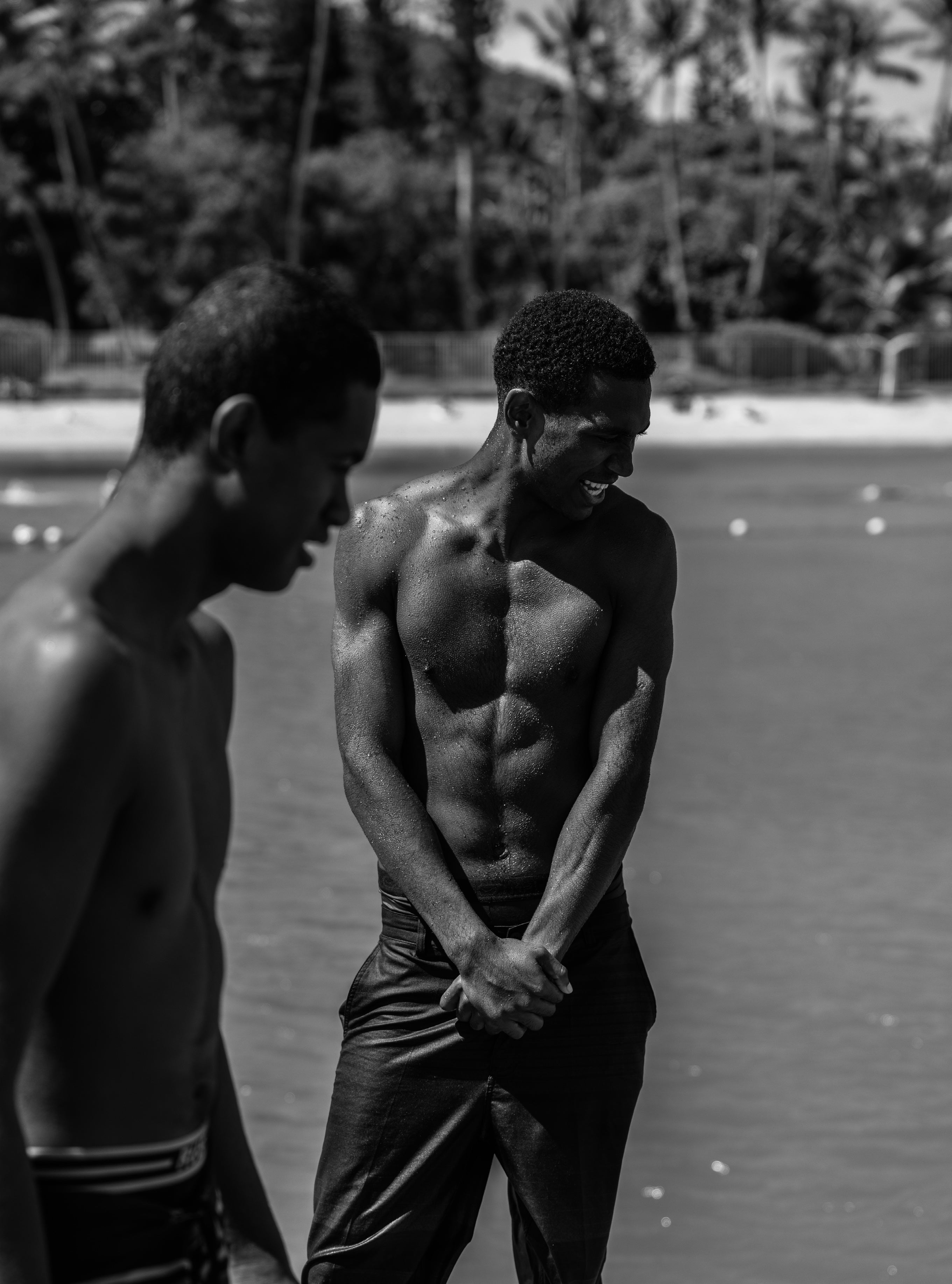 Black and white candid portrait of two young men laughing together by the ocean with palm trees and beach in the background
