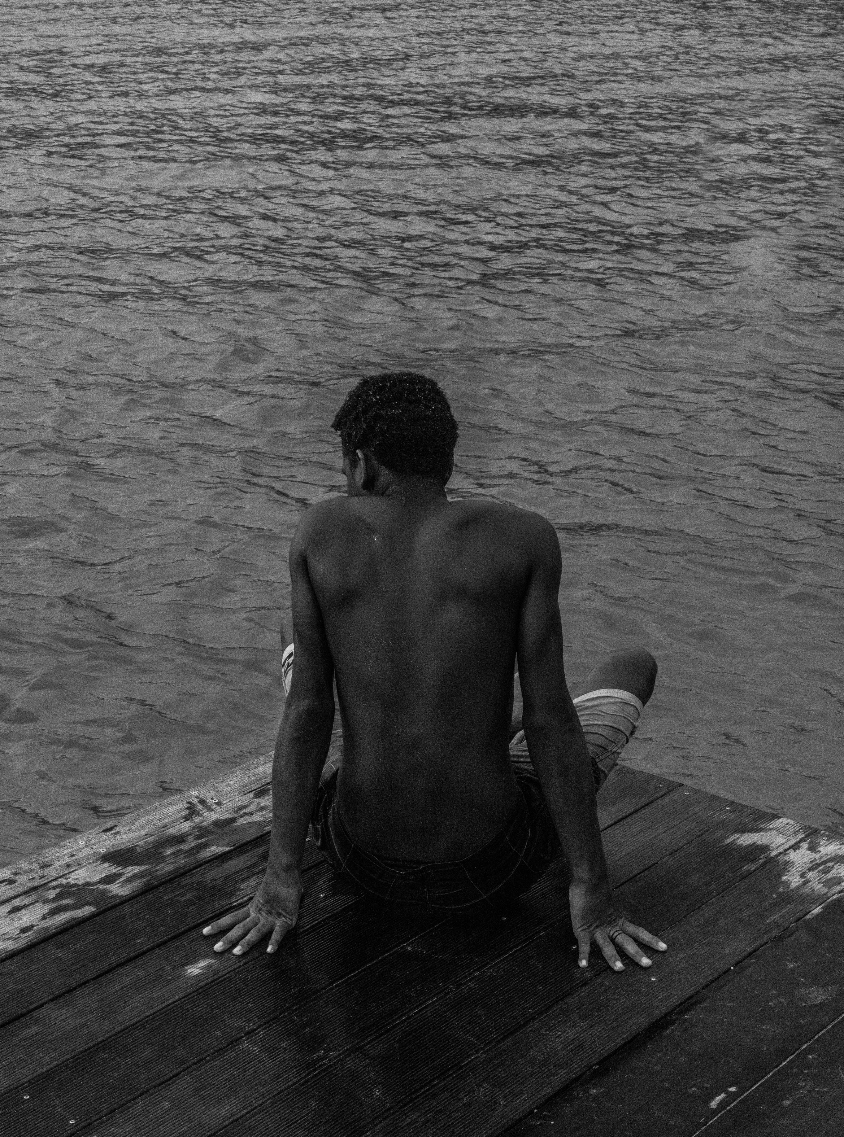 Black and white photograph of a young man sitting at the edge of a wooden dock with his back to camera looking out over rippled water